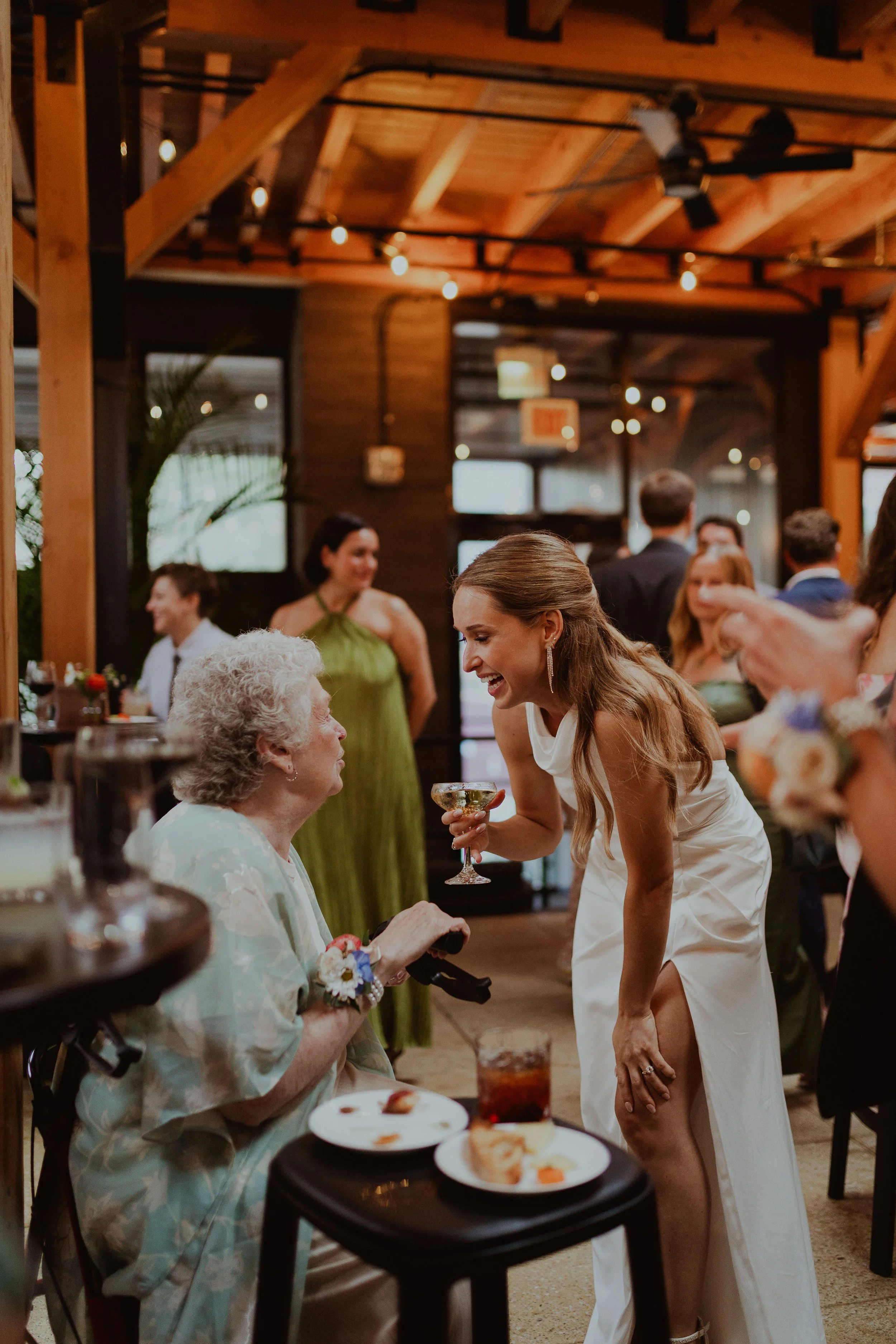 A young bride in a white dress laughing and talking to an elderly woman seated at a table during a celebration or party in a warmly lit, wooden interior setting with other guests in the background. Chicago Winery Wedding photos by Ttolbphotog