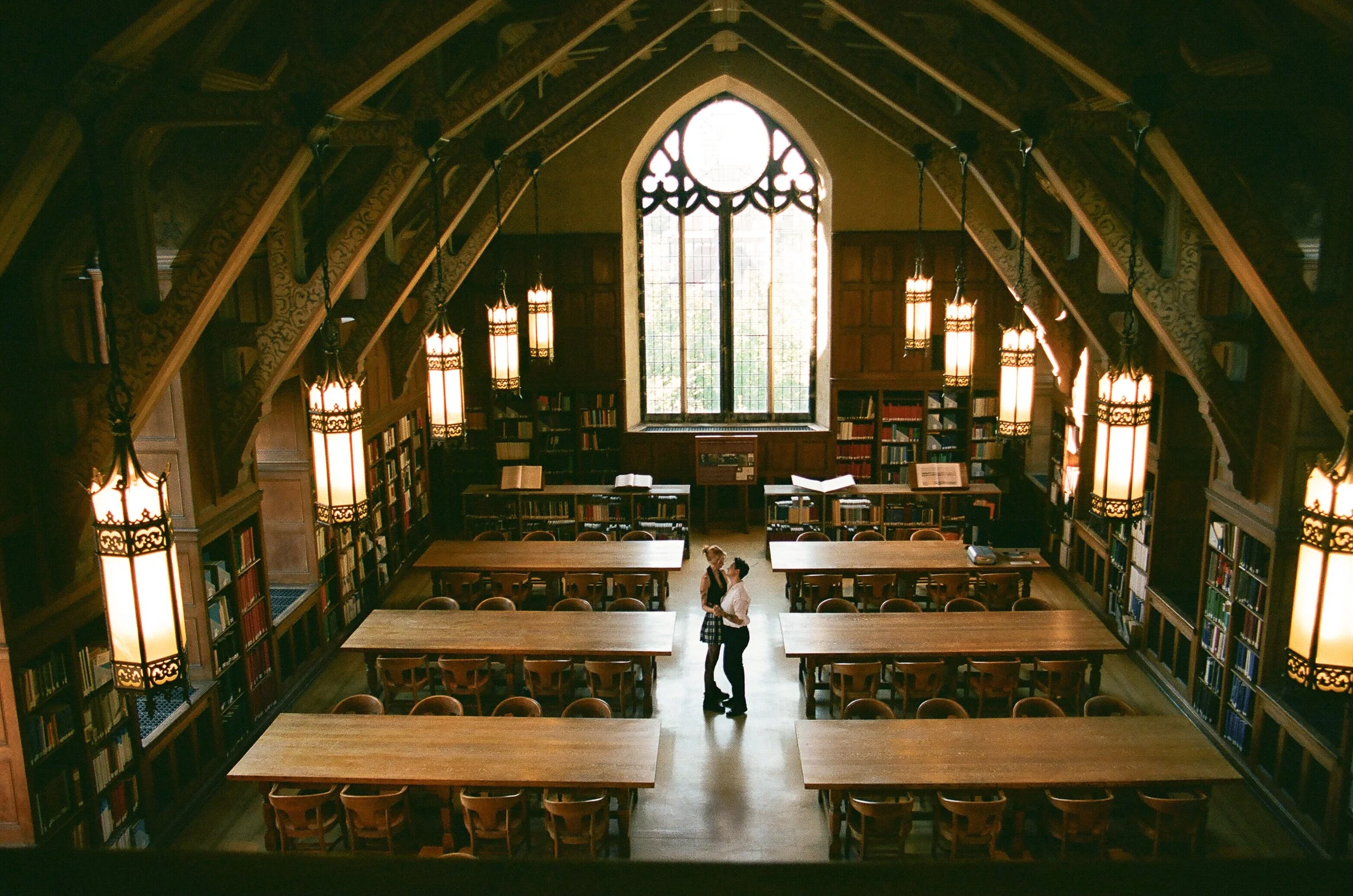 newly engaged couple- both women face each other and talk in a large, old-fashioned library with high wooden ceiling, large arched window, and hanging lamps. Shot on 35mm film by Chicago Photographer Ttolbphotog
