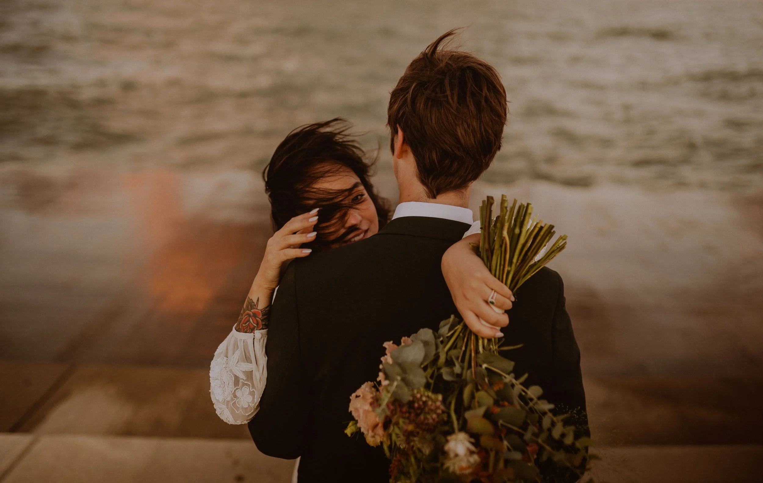 Newlyweds hugging near the lake Michigan, the woman with dark hair and a tattoo on her arm, holding a bouquet of flowers. The man has brown hair and is dressed in a suit, facing away from the camera. Chicago Elopement Photos by Ttolbphotog