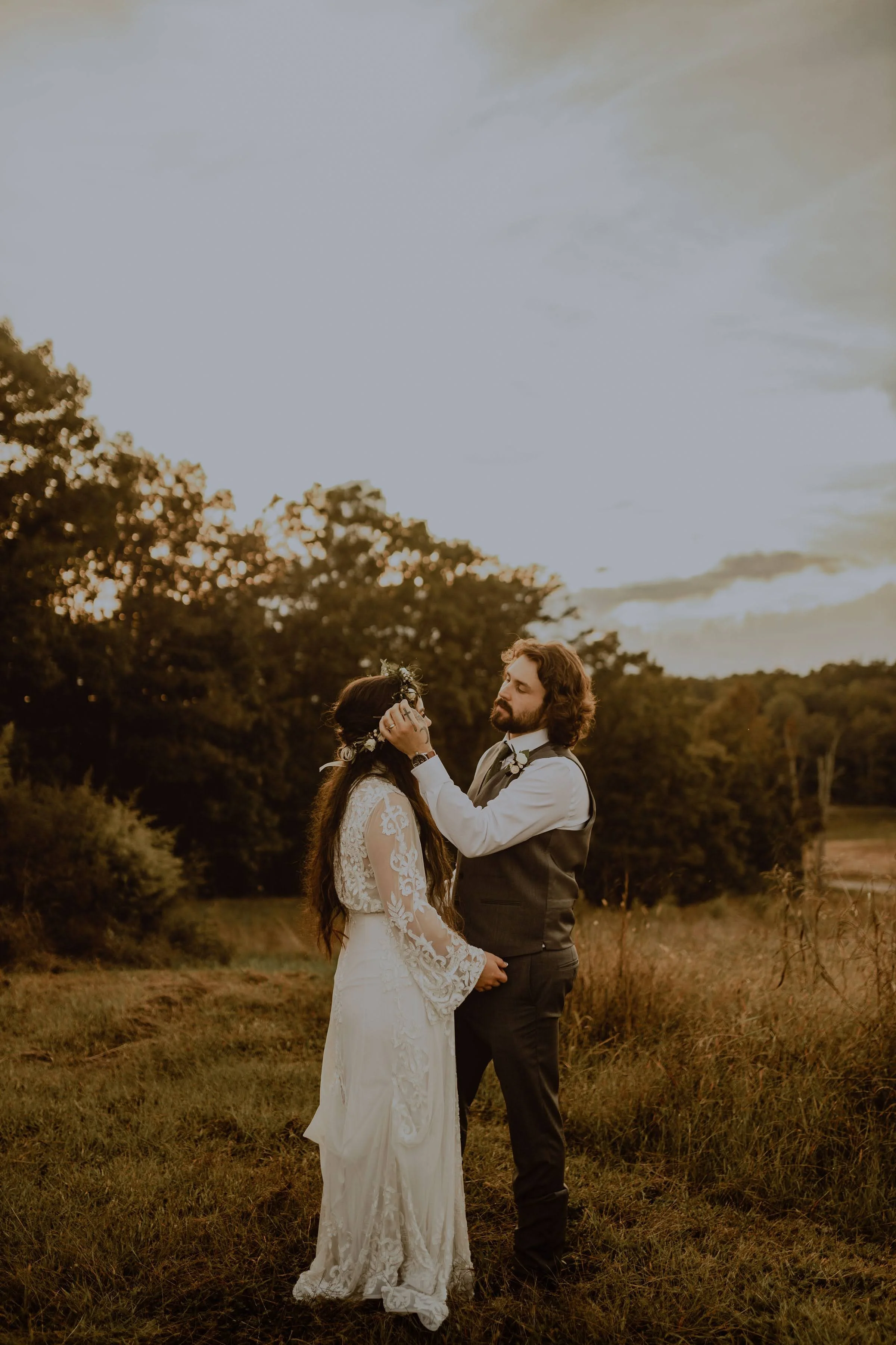 A groom places a floral crown on a bride's head during a rustic outdoor wedding at sunset.