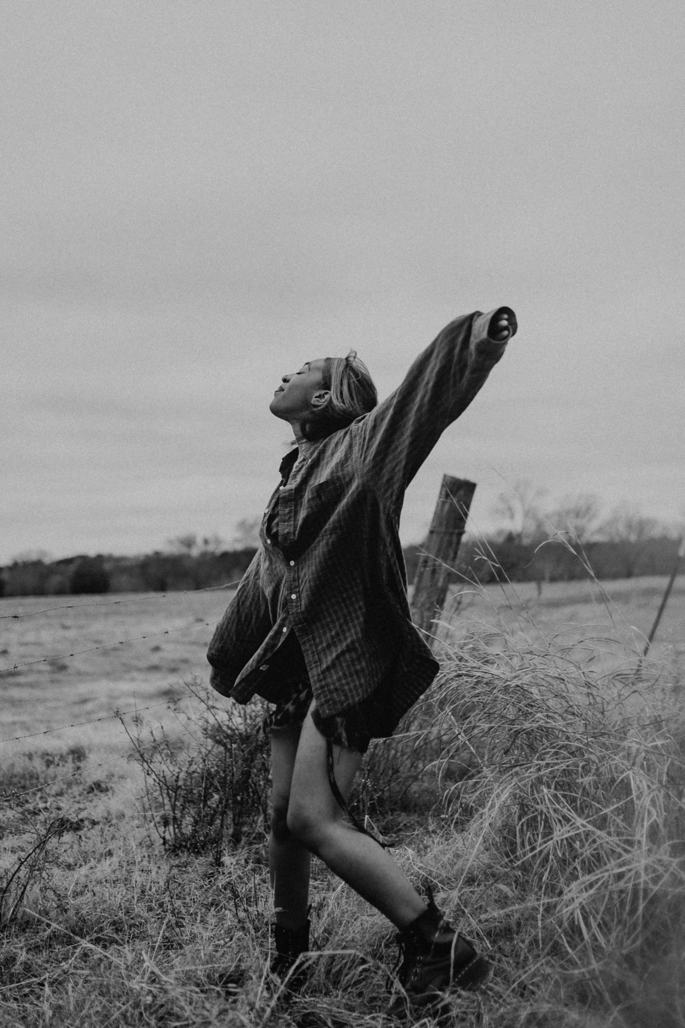A black and white photo of a woman standing outdoors in a field, with one arm raised and her head tilted back, wearing a loose jacket, shorts, and boots.