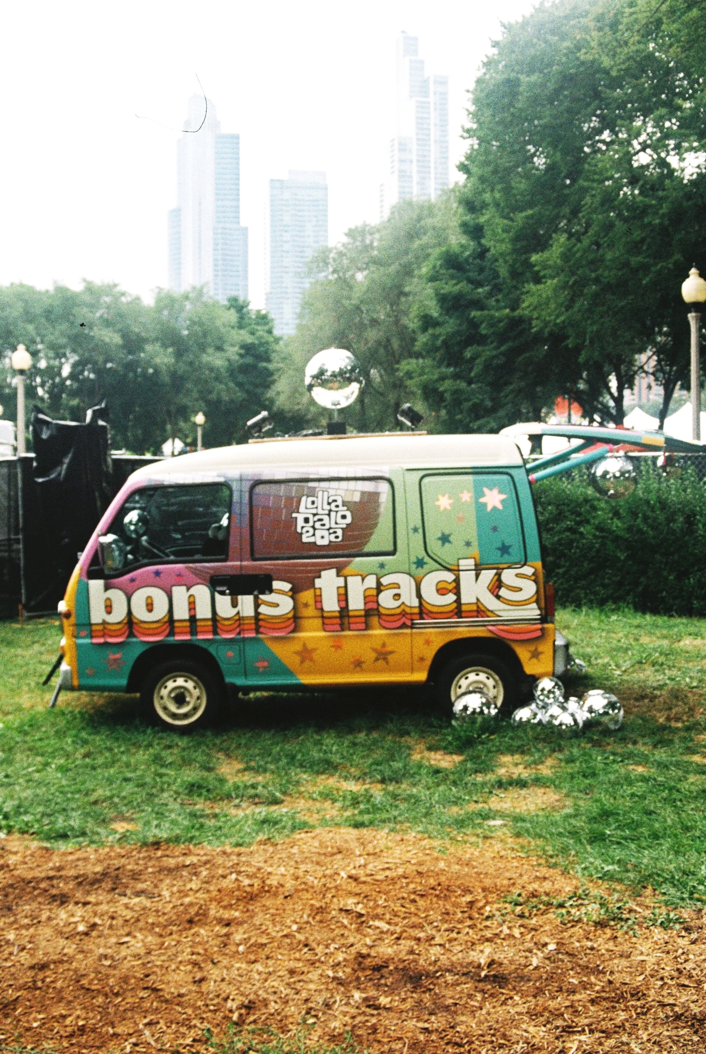 image of a vintage bus with "lollapalooza BONUS TRACKS" on the side- COlorful van is seen on the grass with the Chicago skyline peeking above the trees of the park at Lollapalooza- image by Taylor with TTOLBPHOTOG chicago photographer