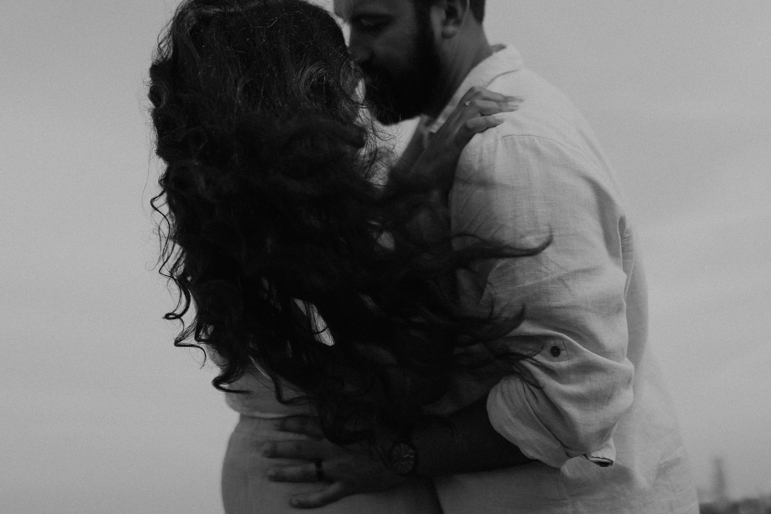 A black-and-white photo of a couple embracing closely, with the woman having long, curly hair and the man with a beard, both with their eyes closed.