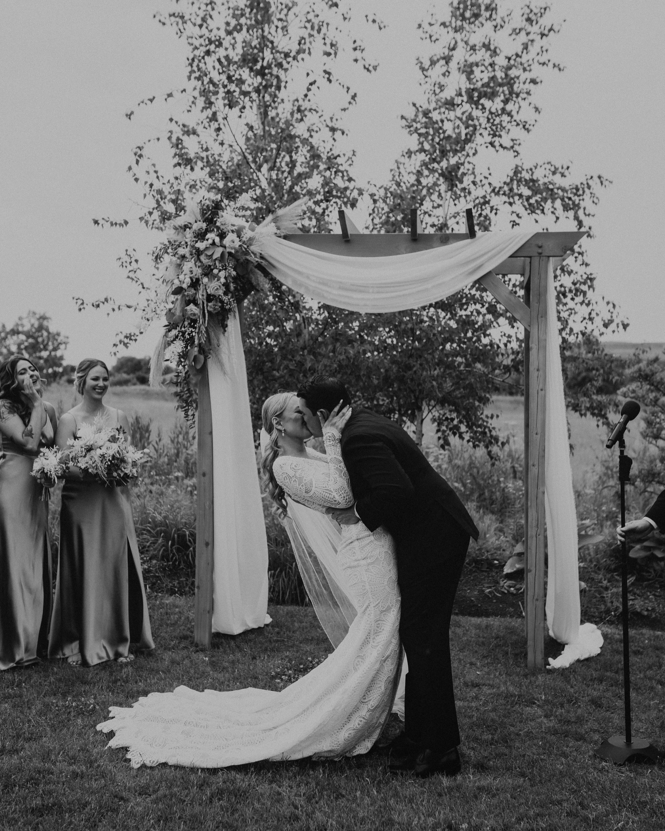 A black and white photo of a bride and groom kissing under an outdoor wooden arch decorated with flowers and fabric, with bridesmaids standing nearby.