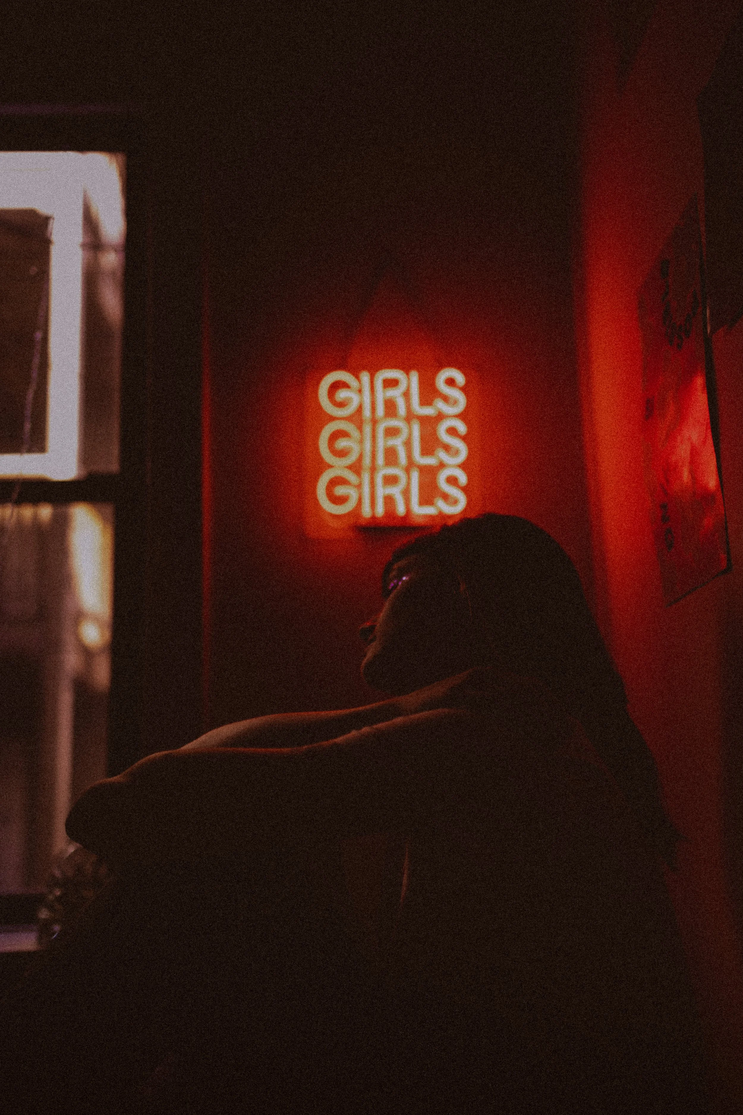 A woman sitting in a dimly lit room with a red neon sign that says 'GIRLS' on the wall behind her.
