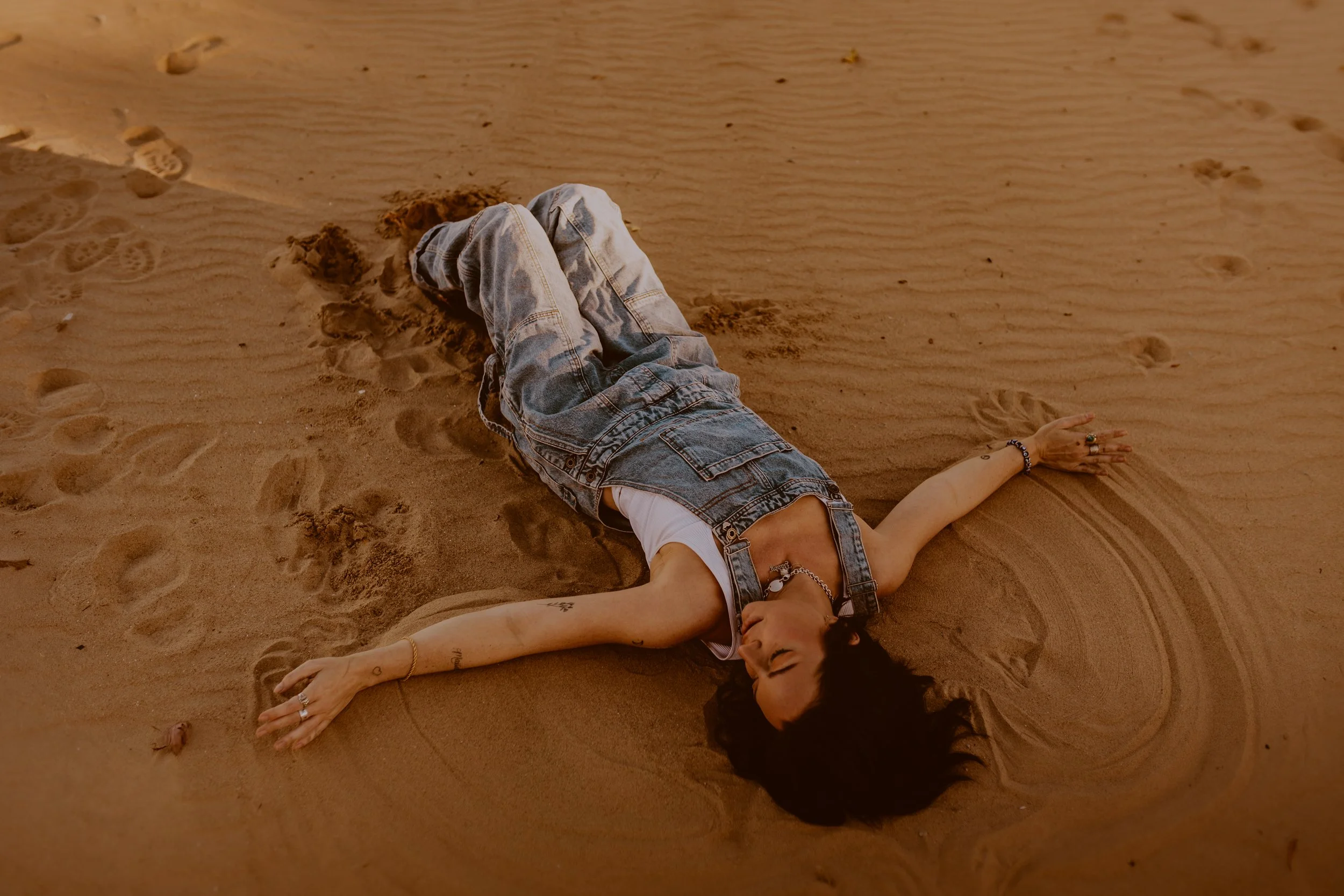 Woman lying on sand at Osterman Beach in Lincoln Park Chicago. She is wearing denim overalls with a white tank top underneath various tattoos showing, with her eyes closed and surrounded by footprints.