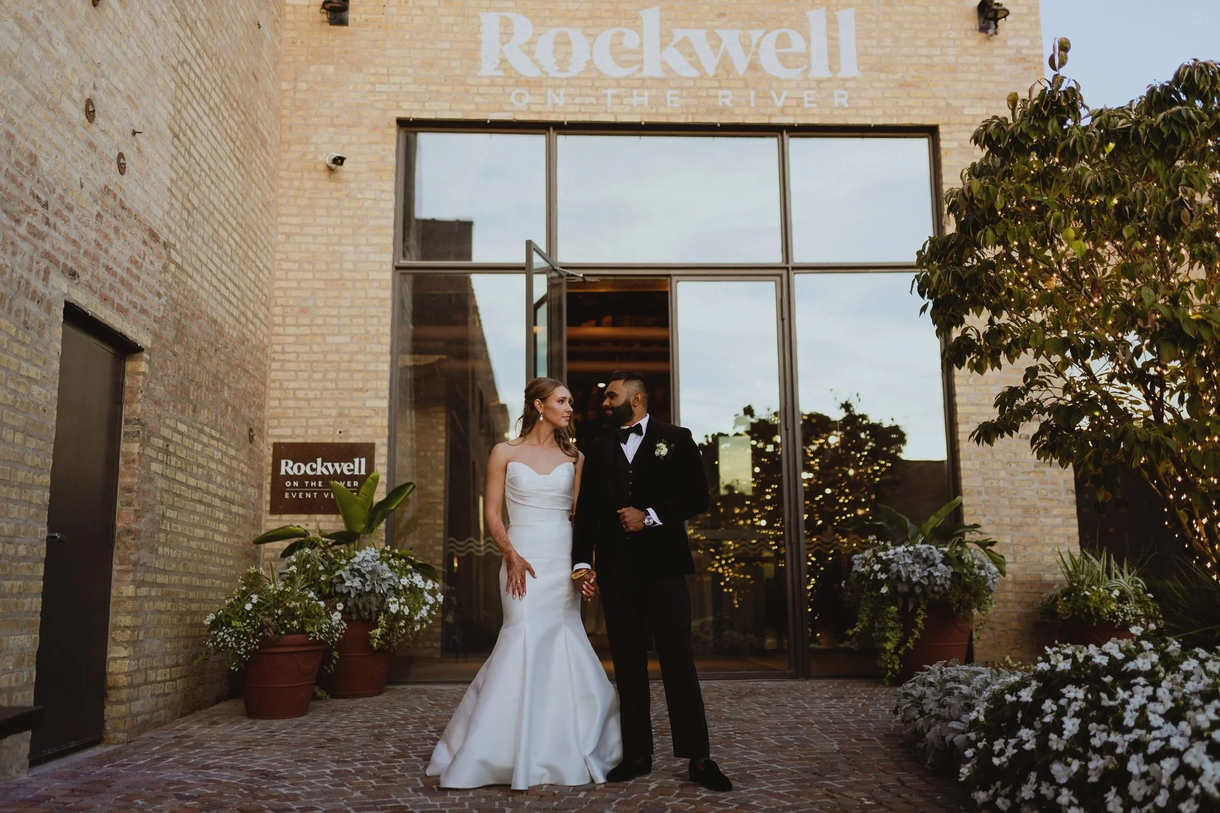 A bride and groom standing outside of Rockwell on the River, holding hands and looking at each other, surrounded by potted plants and white flowers.