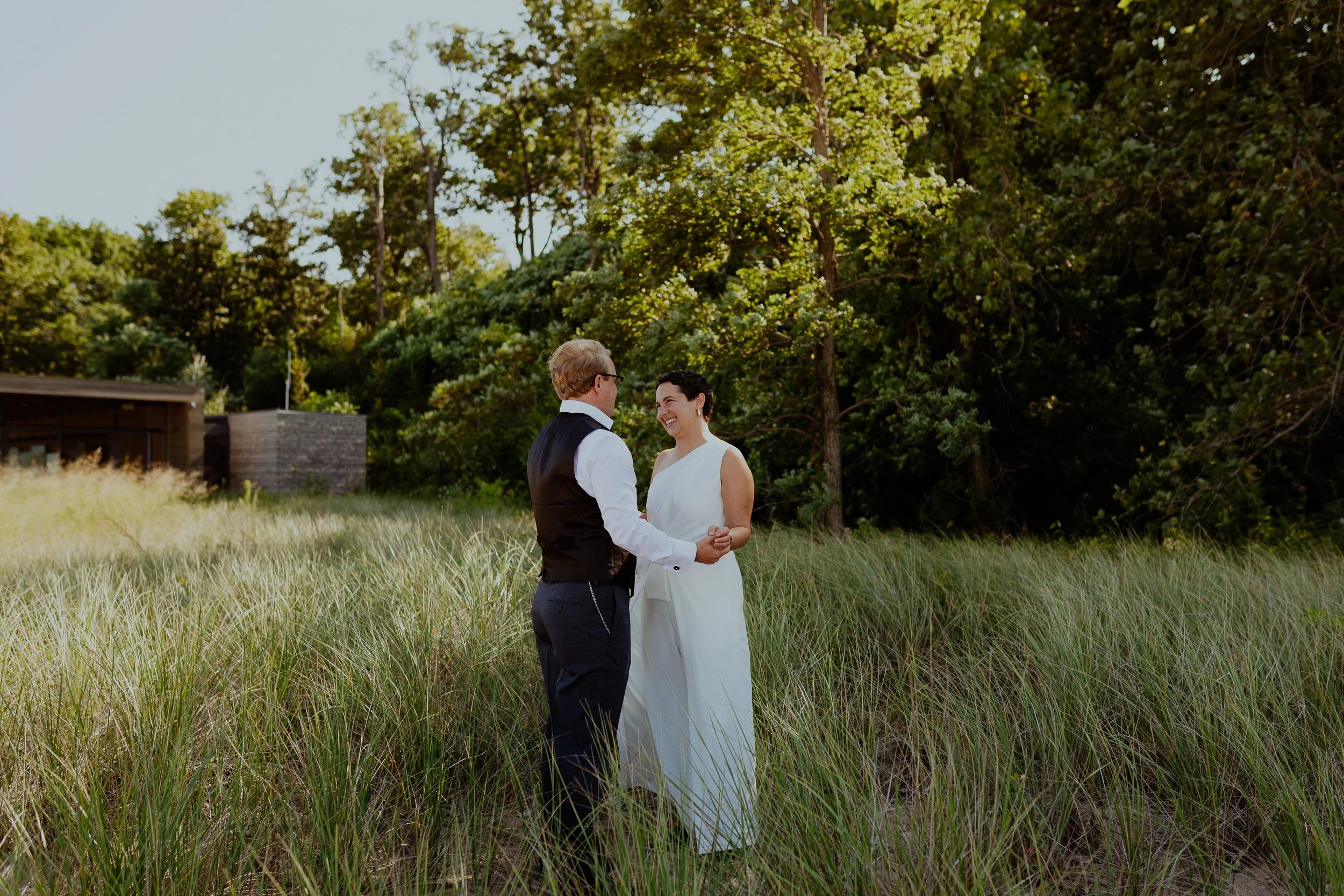 A couple holding hands and smiling at each other outdoors in a grassy field with trees in the background during daytime.