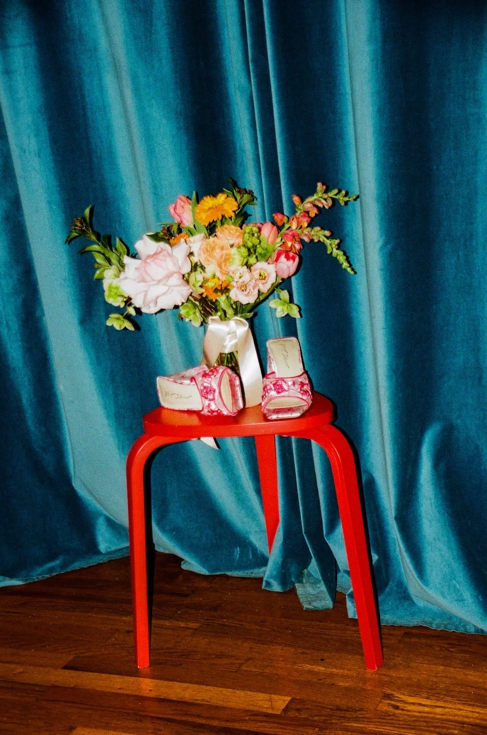 A red stool with a vase of pink, white, and orange flowers and a pair of pink floral high-heeled shoes on top, set against a blue velvet curtain background. Film photography Intimate Micro Wedding Photos by ttolbphotog Logan Square Chicago