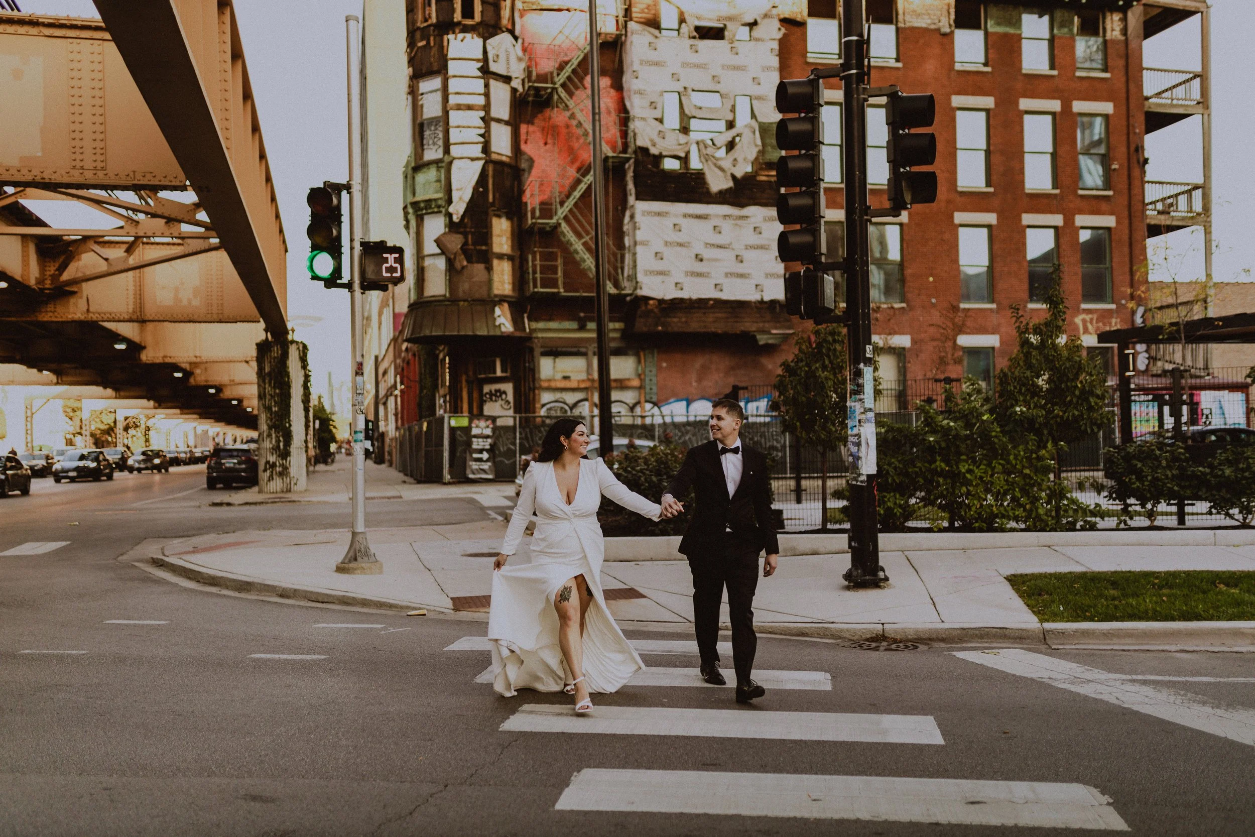 A couple in wedding attire crossing a city street at a crosswalk during sunset. The woman is in a white wedding dress with a tattoo on her thigh, and the man is in a black tuxedo. There are traffic lights and urban buildings in the background.