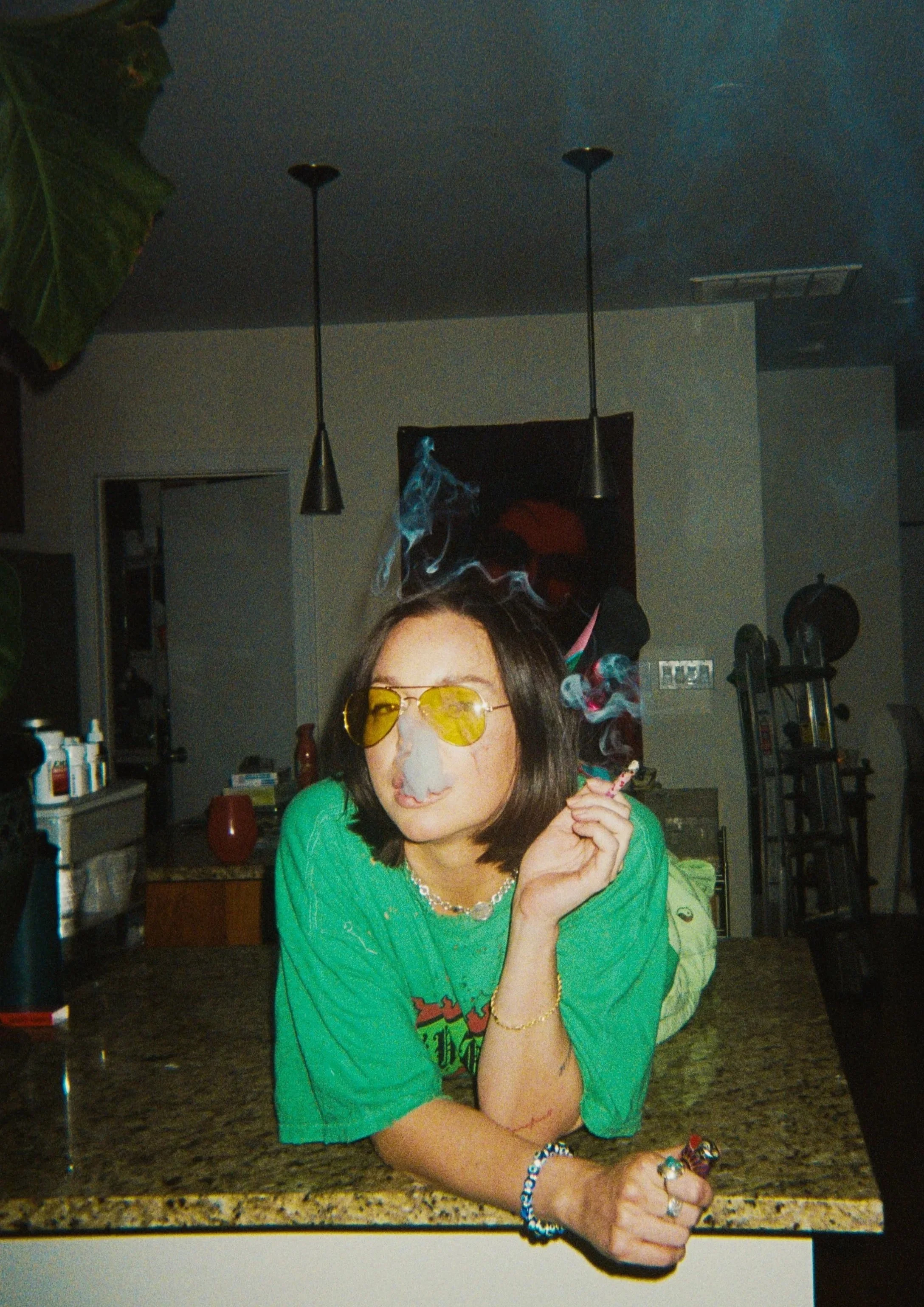 A woman with shoulder-length dark hair wearing yellow sunglasses, a green T-shirt, and jewelry, is leaning on a kitchen counter, smoking a cigarette with smoke swirling around her face.