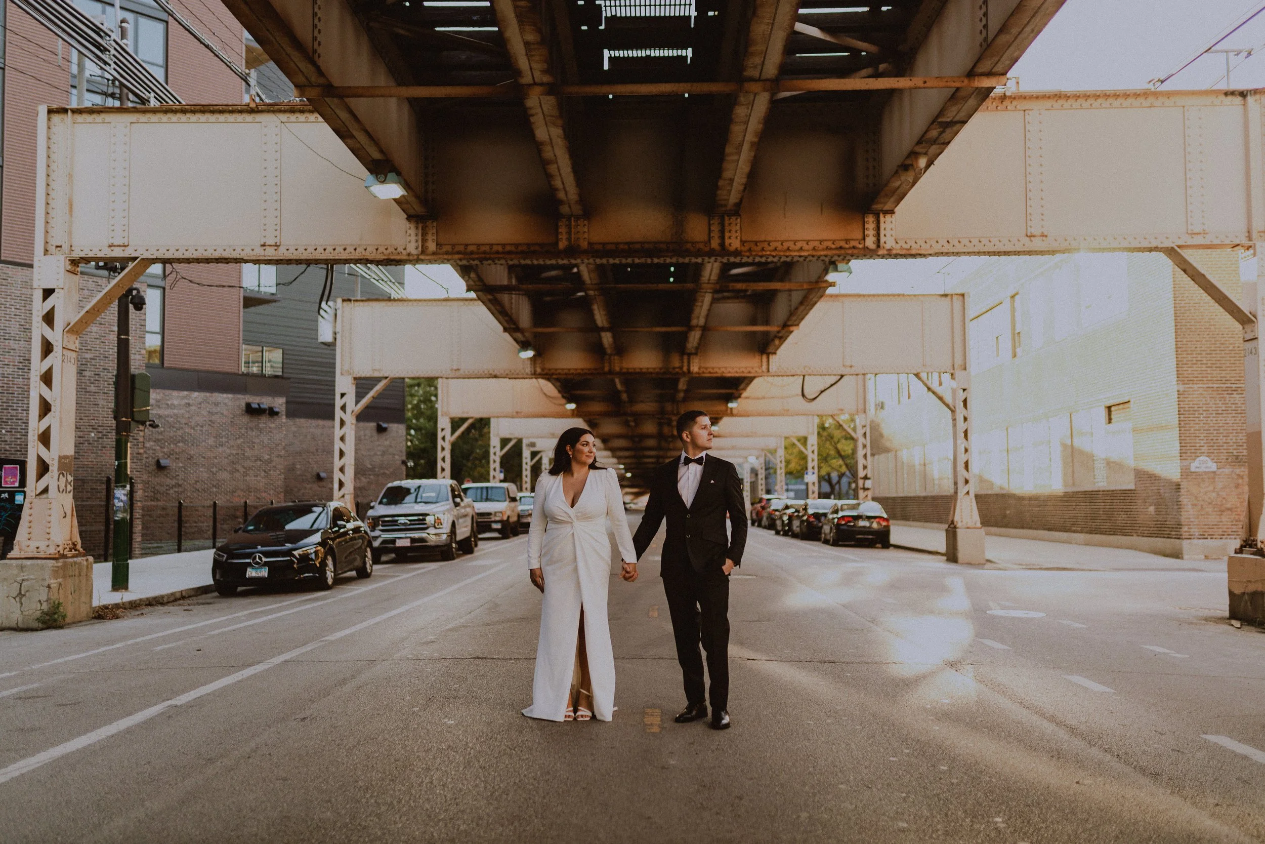 A couple in wedding attire walking hand in hand under an elevated train track on an urban street, with parked cars and brick buildings in the background.