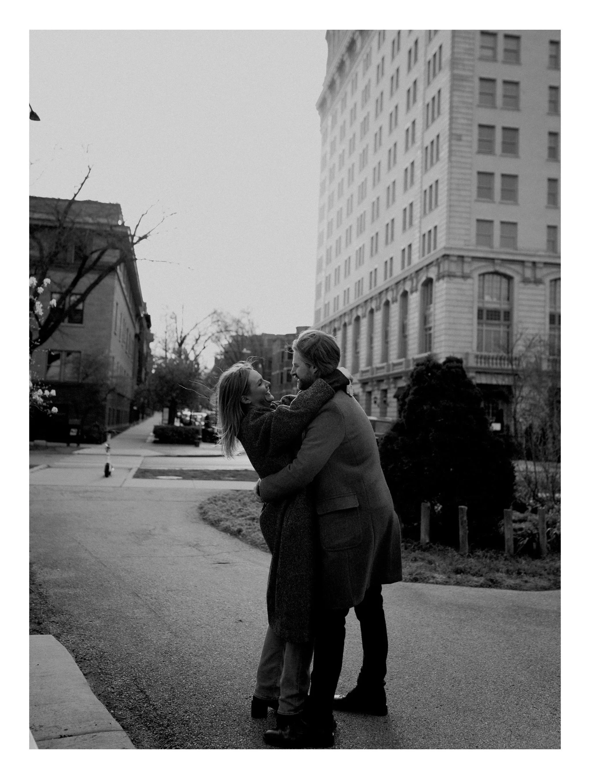 A couple embracing and smiling outdoors at Lincoln Park garden with tall buildings and trees in the background, black and white photo.