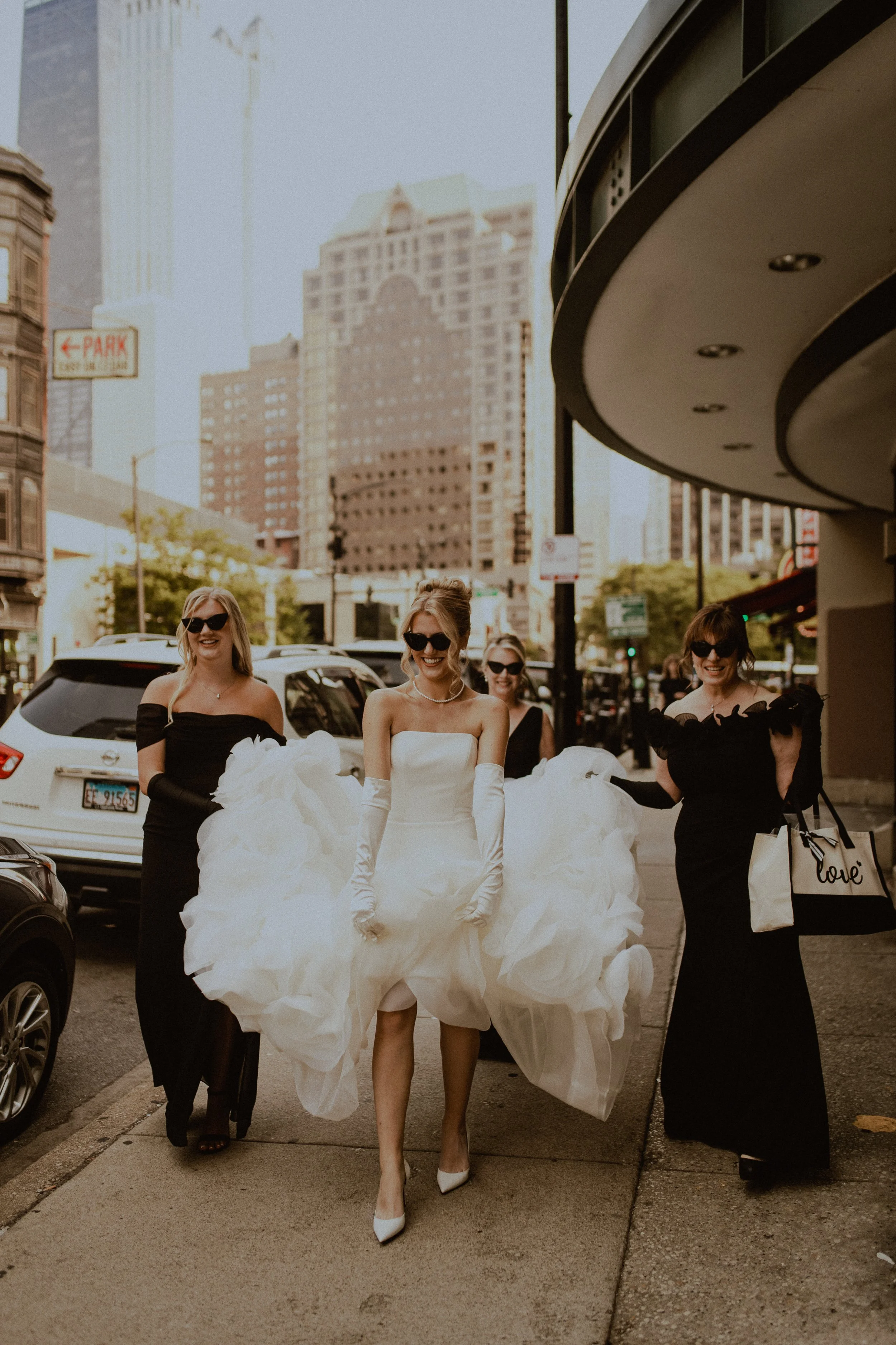 A bride in a white wedding gown and long gloves walking on an urban sidewalk with four women in black dresses, all wearing sunglasses, in a city street with tall buildings and cars in the background.