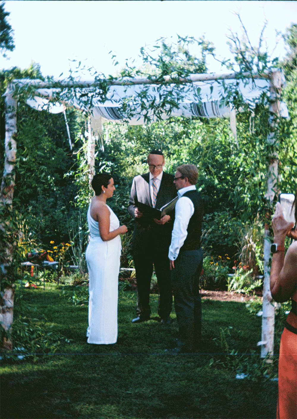 A couple getting married during an outdoor jewish ceremony under a decorated white archway with greenery, with an officiant present and guests taking photos. Documentary Wedding photos taken by Ttolbphotog