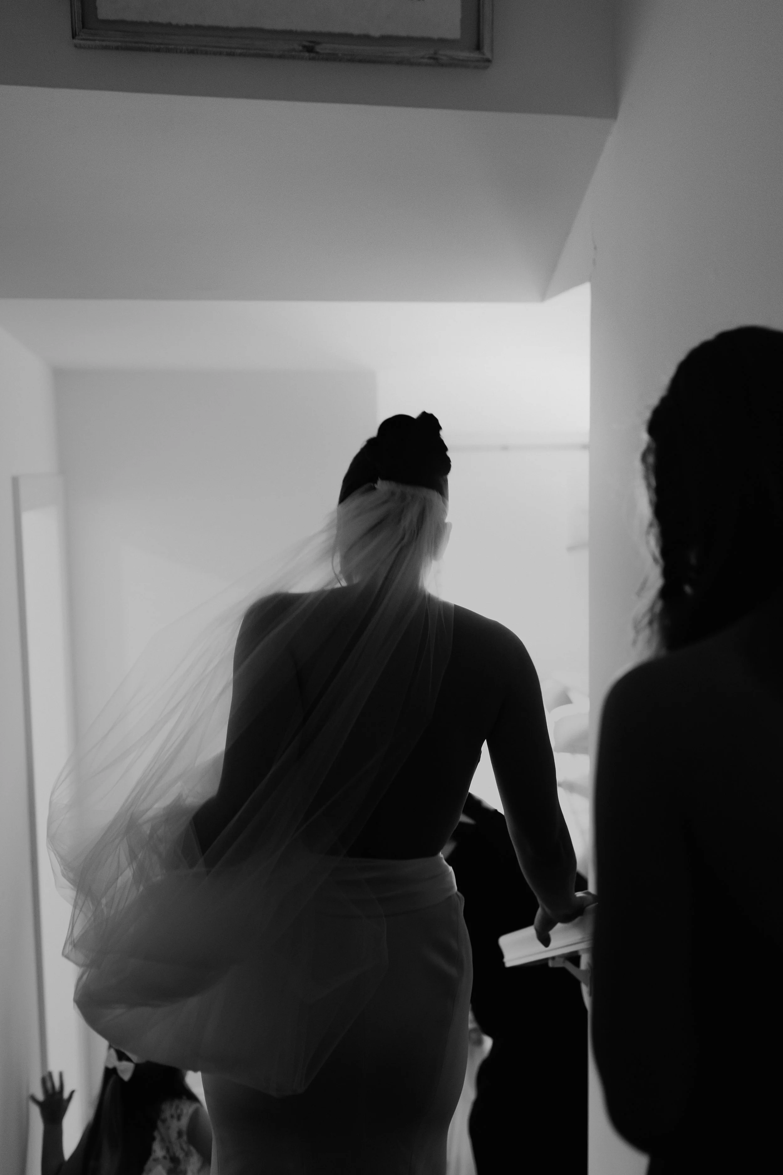 Black and white photo of a woman wearing a veil, facing away, with a wedding dress and a veil flowing over her shoulder. There are children in the background, one with hands raised, in a room with plain walls and a ceiling picture frame.