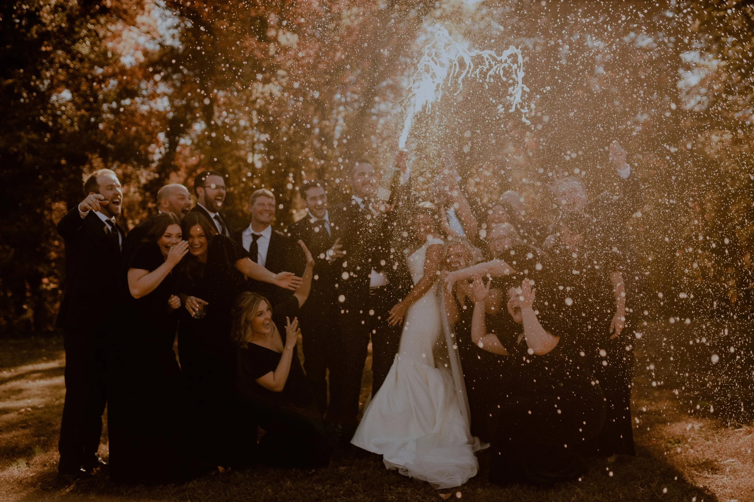 A group of people celebrating outdoors with champagne, some wearing suits and a wedding dress, at sunset during a wedding celebration.