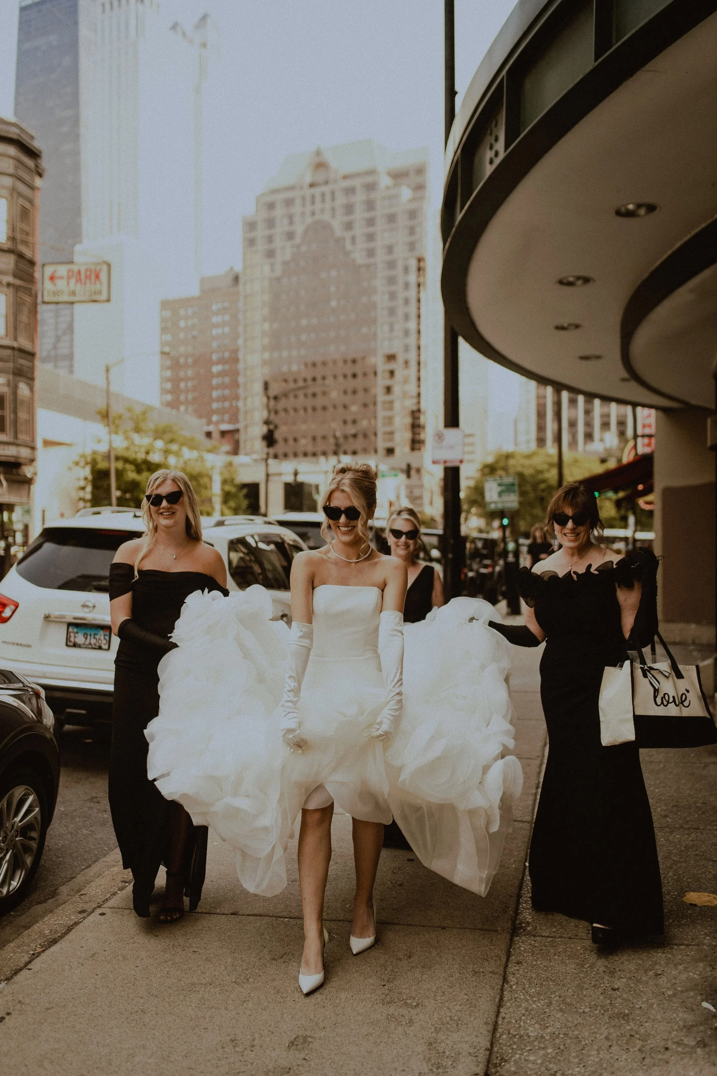 A bride in a white wedding dress walking on a Chicago city sidewalk with three women dressed in black. The women are smiling and wearing sunglasses. The background shows tall buildings, cars parked on the street, and a parking sign. Wedding photos