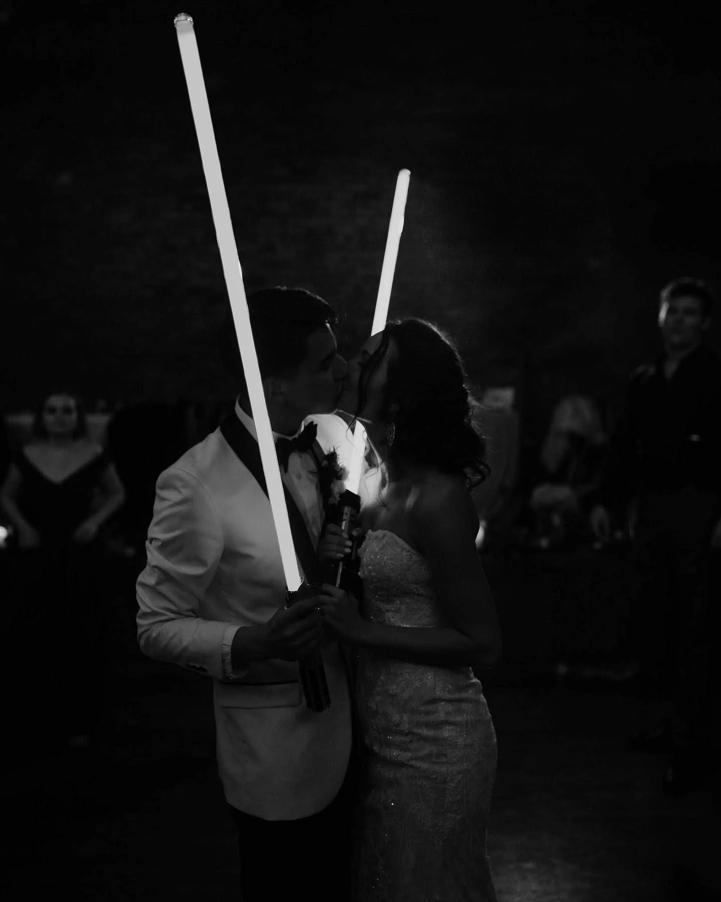 A black and white photo of a couple kissing during a wedding reception, holding illuminated sticks, with guests in the background.