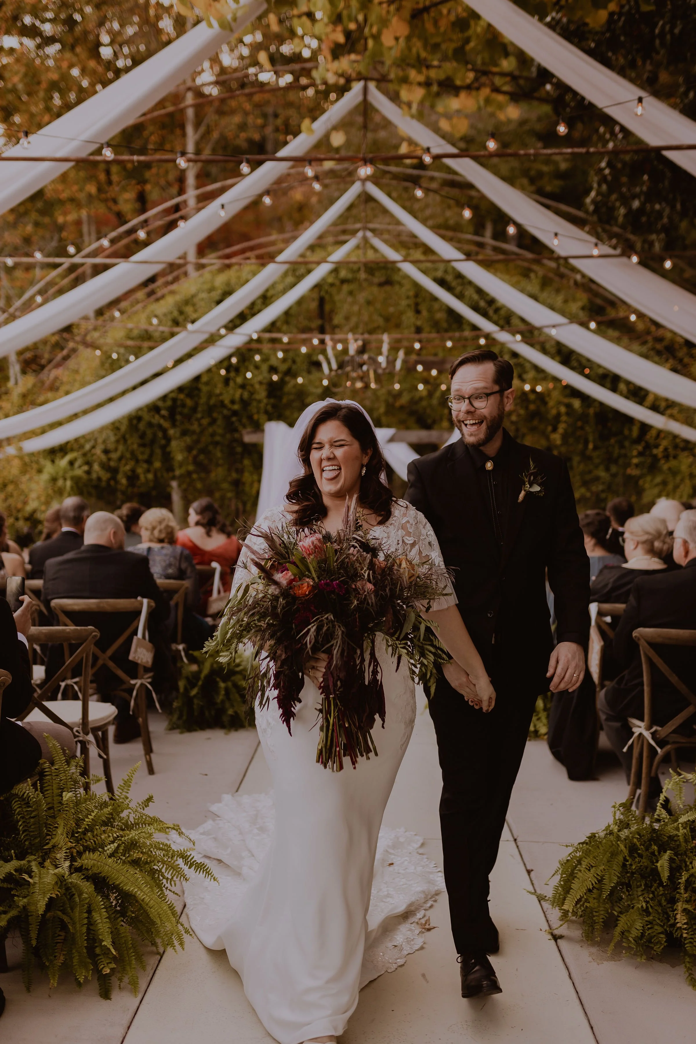 A bride and groom walk down the aisle during their outdoor wedding ceremony at sunset. The bride is wearing a white dress and holding a large bouquet, and the groom is in a black suit. Guests are seated on either side under string lights and a decora