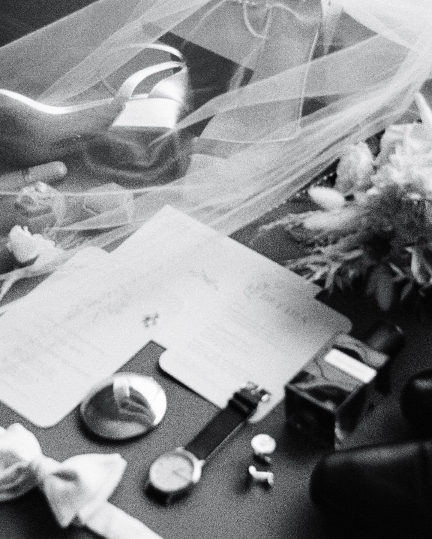A desk surface with a wedding or event invitation, a wristwatch, a small box, and scattered flowers, along with some other small items and a plastic bag containing a shoe.