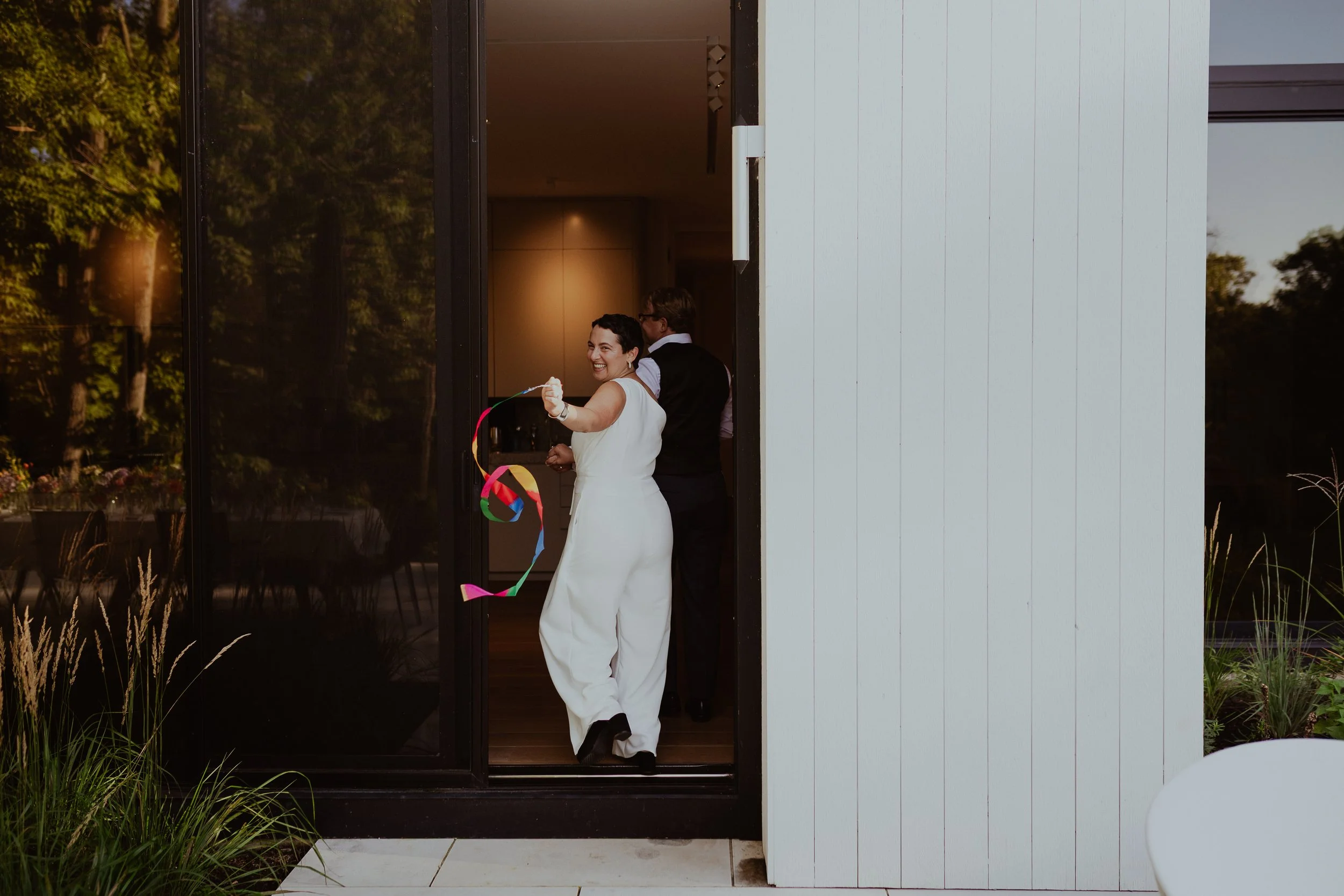 A woman in a white jumpsuit holding a colorful ribbon, smiling, as she steps out of a modern house with glass doors and white siding, with a man in a black vest and white shirt behind her.