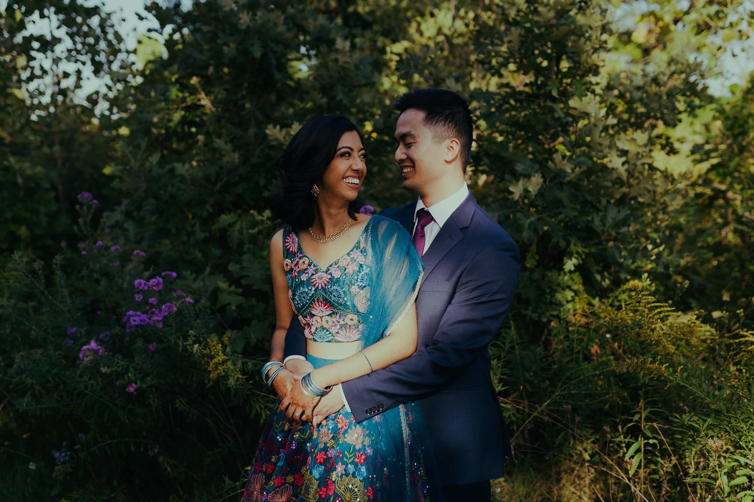 A joyful couple, dressed in formal attire, standing outdoors amid green foliage and purple flowers, smiling at each other.