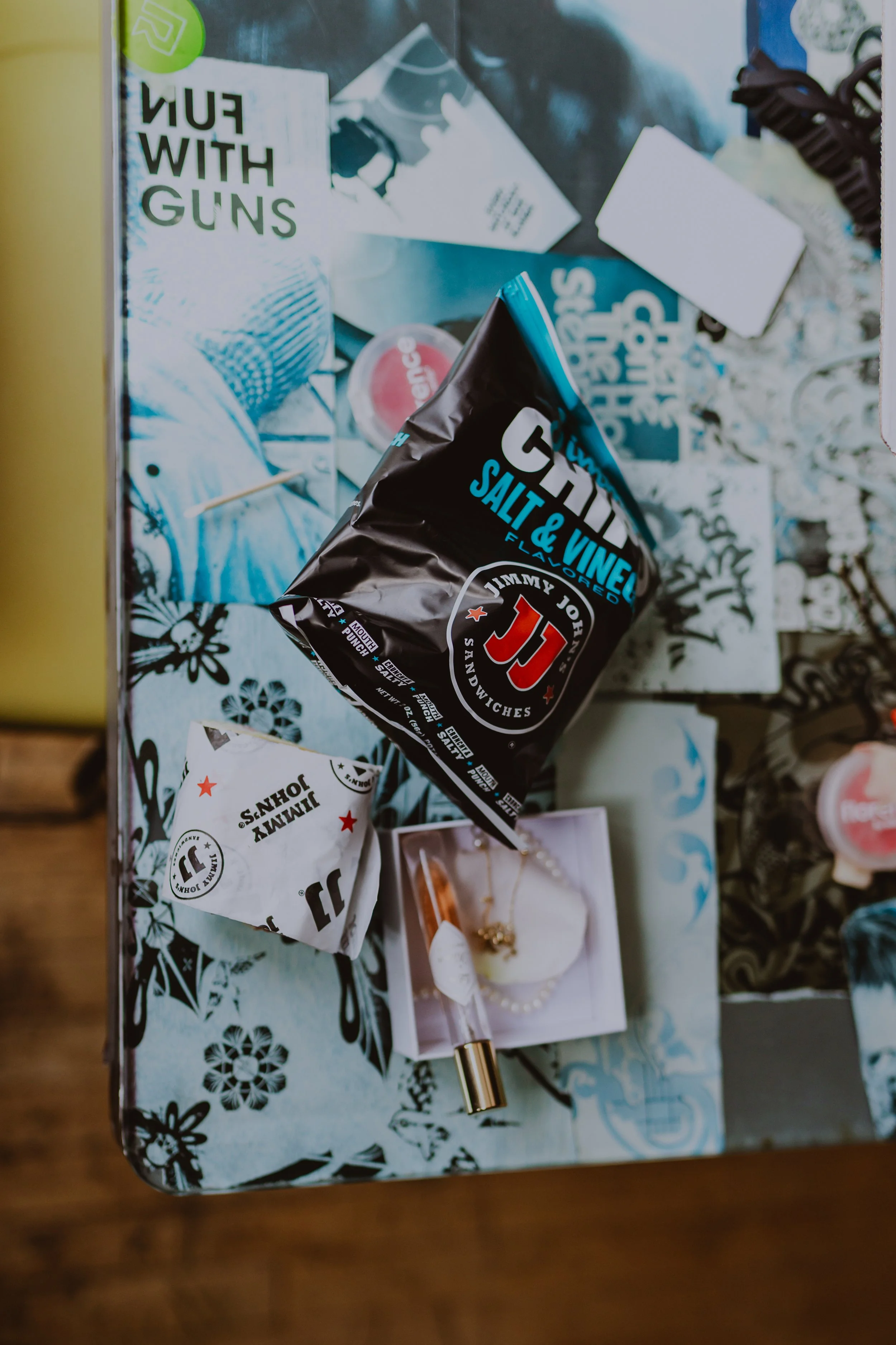 Top view of a cluttered surface with a Jimmy John's sandwich wrap, a salt and vinegar flavored chip bag, a jewelry box with a necklace and lip gloss, and various miscellaneous items including a magazine and a card.