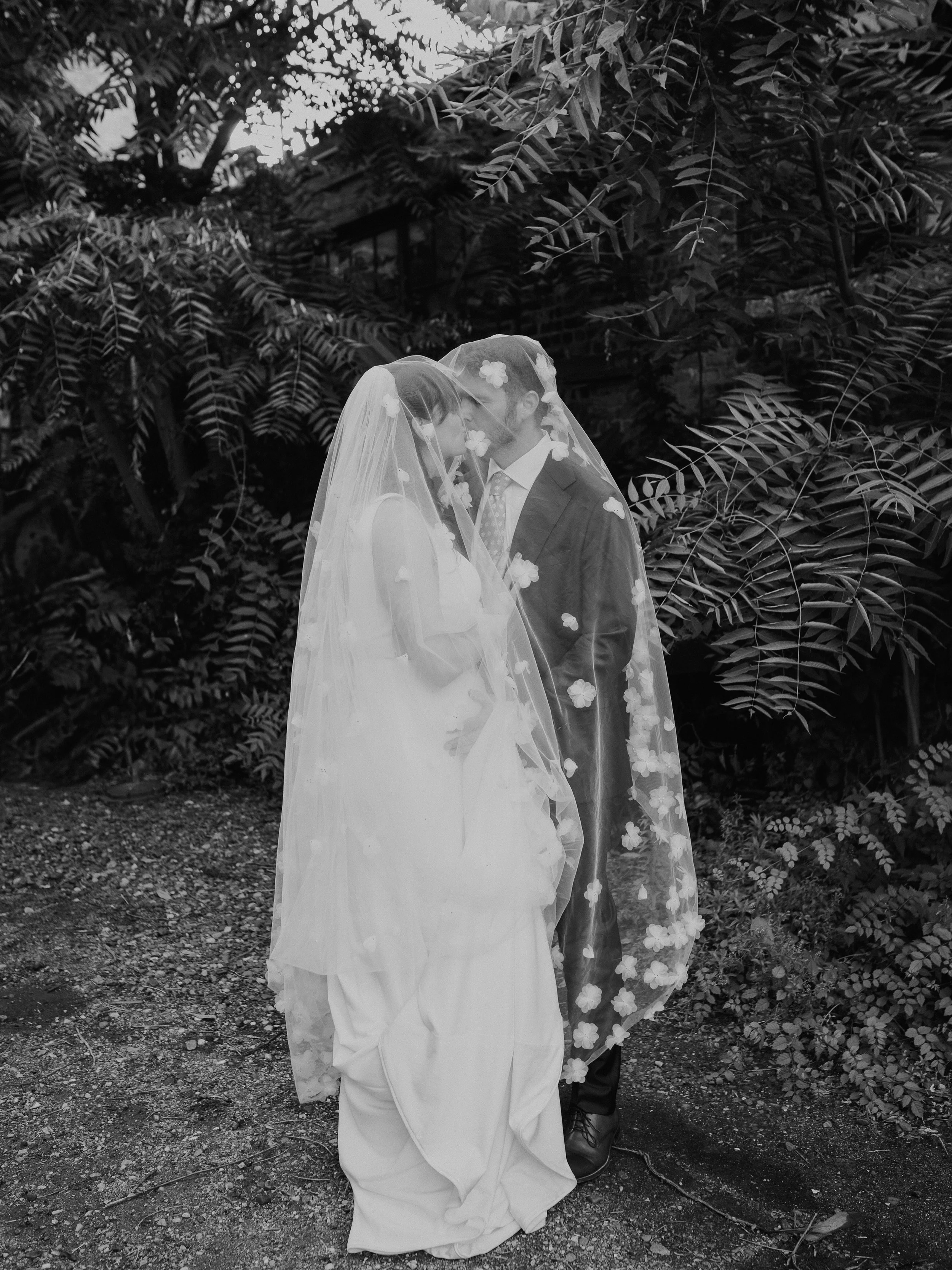 A black and white photo of a bride and groom standing close together, sharing a kiss beneath a sheer veil decorated with floral patterns, outdoors with lush foliage in the background. Salvage One Wedding Venue Chicago IL