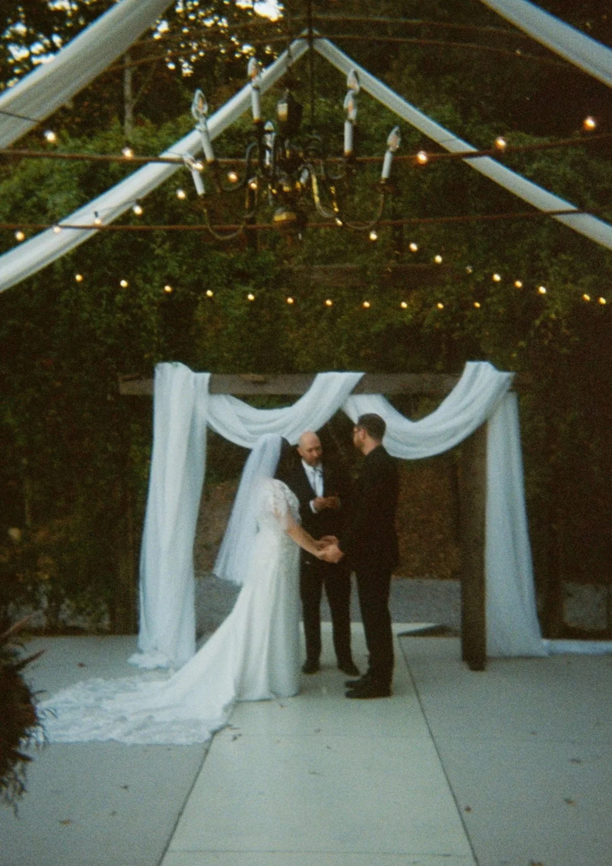 A couple getting married under a decorated archway, holding hands and facing each other, with two officiants standing behind them. The backdrop features trees and draped white fabric. Documentary 35mm Film Wedding Photos.