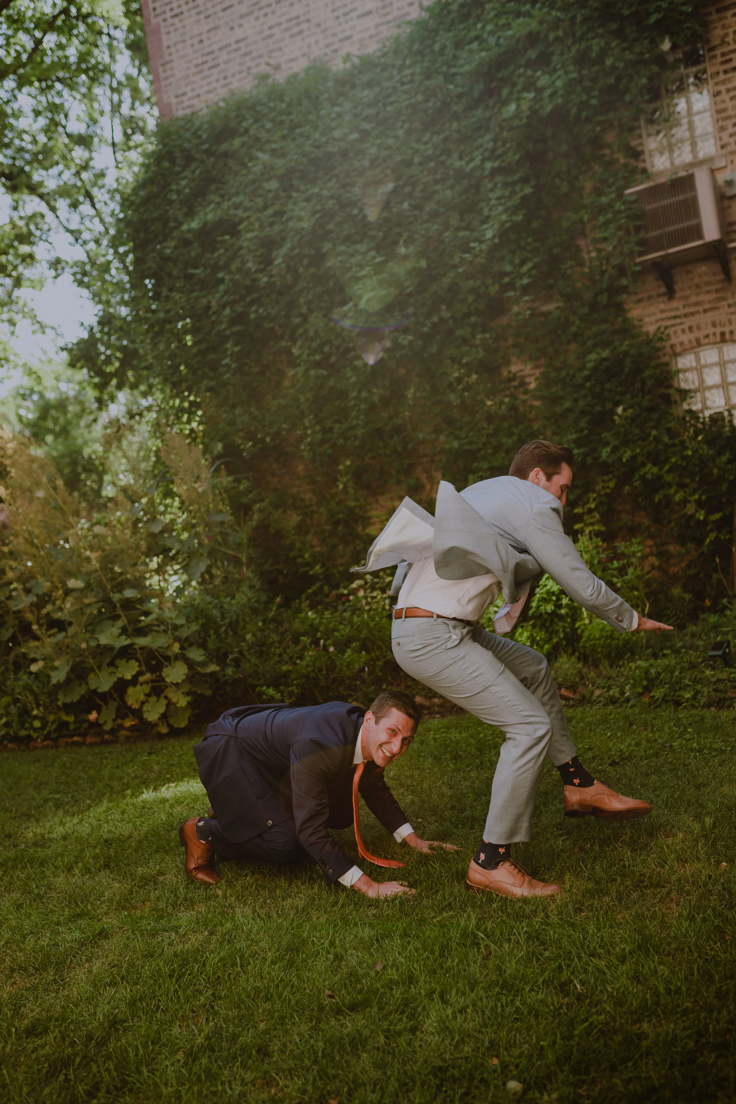 Two men in wedding suits playing leap frog on a grassy lawn. One man is crawling on hands and knees, and the other is in mid-air as if jumping over or avoiding the crawling man. The background features a brick building partially covered in greenery.