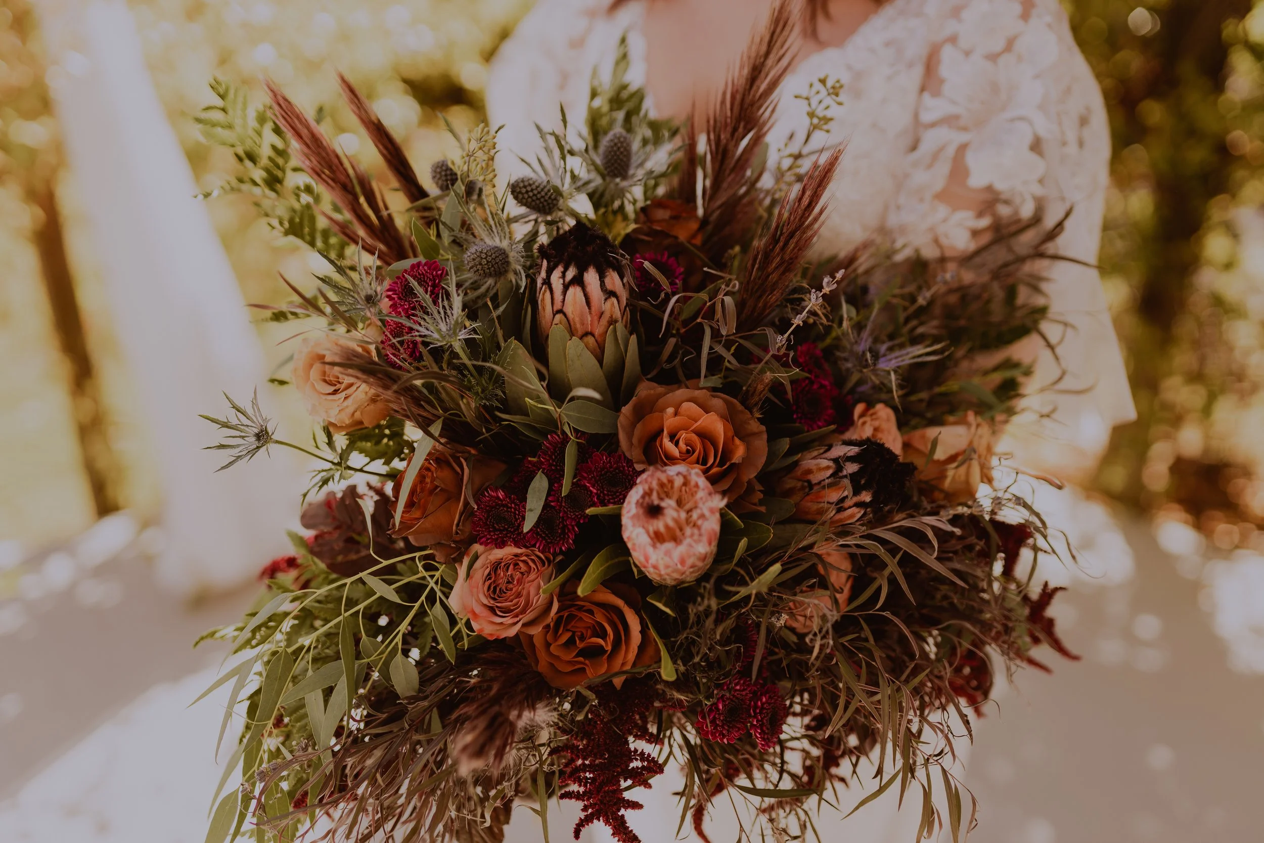 A person holding a large, autumn-themed bouquet of flowers and dried plants, with a blurred background of trees and sunlight.