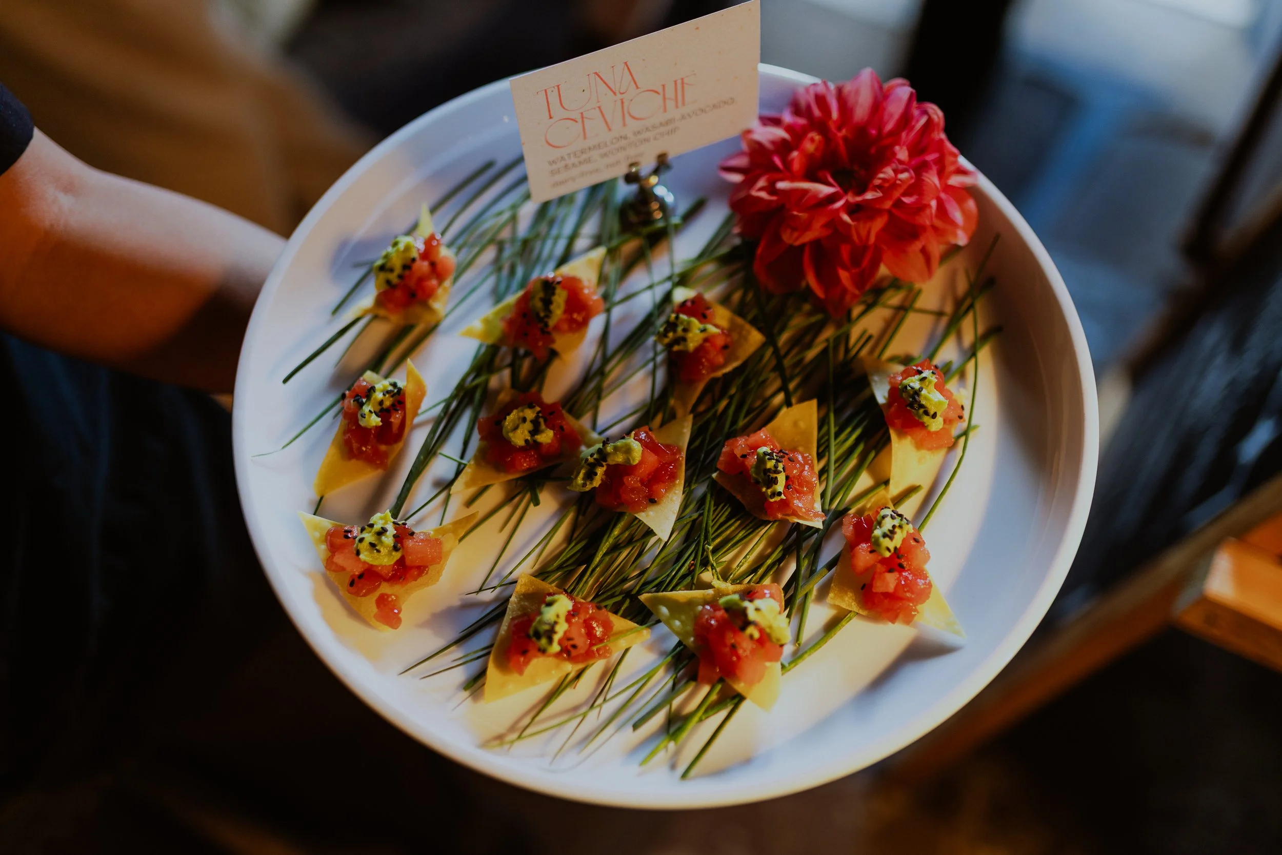 A person holding a white platter with tiny appetizer bites topped with diced tomatoes and black seeds, arranged with chives and garnished with a pink flower and a small sign reading 'Tuna Ceviche'.