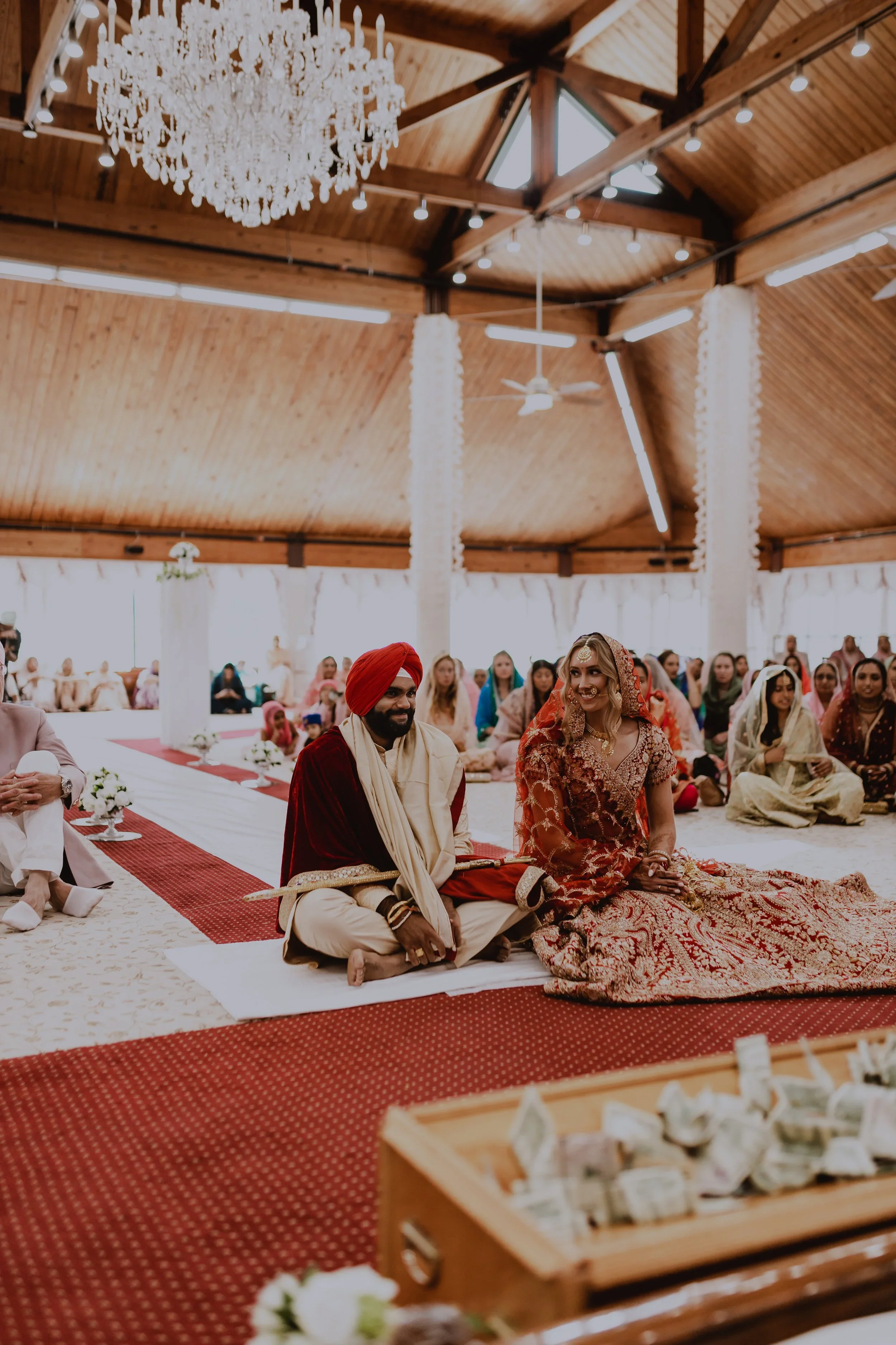 A traditional Indian wedding ceremony inside a wooden pavilion with a high ceiling and chandeliers. The bride and groom sit on the floor on a decorated aisle, surrounded by guests. The groom wears a cream-colored outfit with a red turban and shawl, w