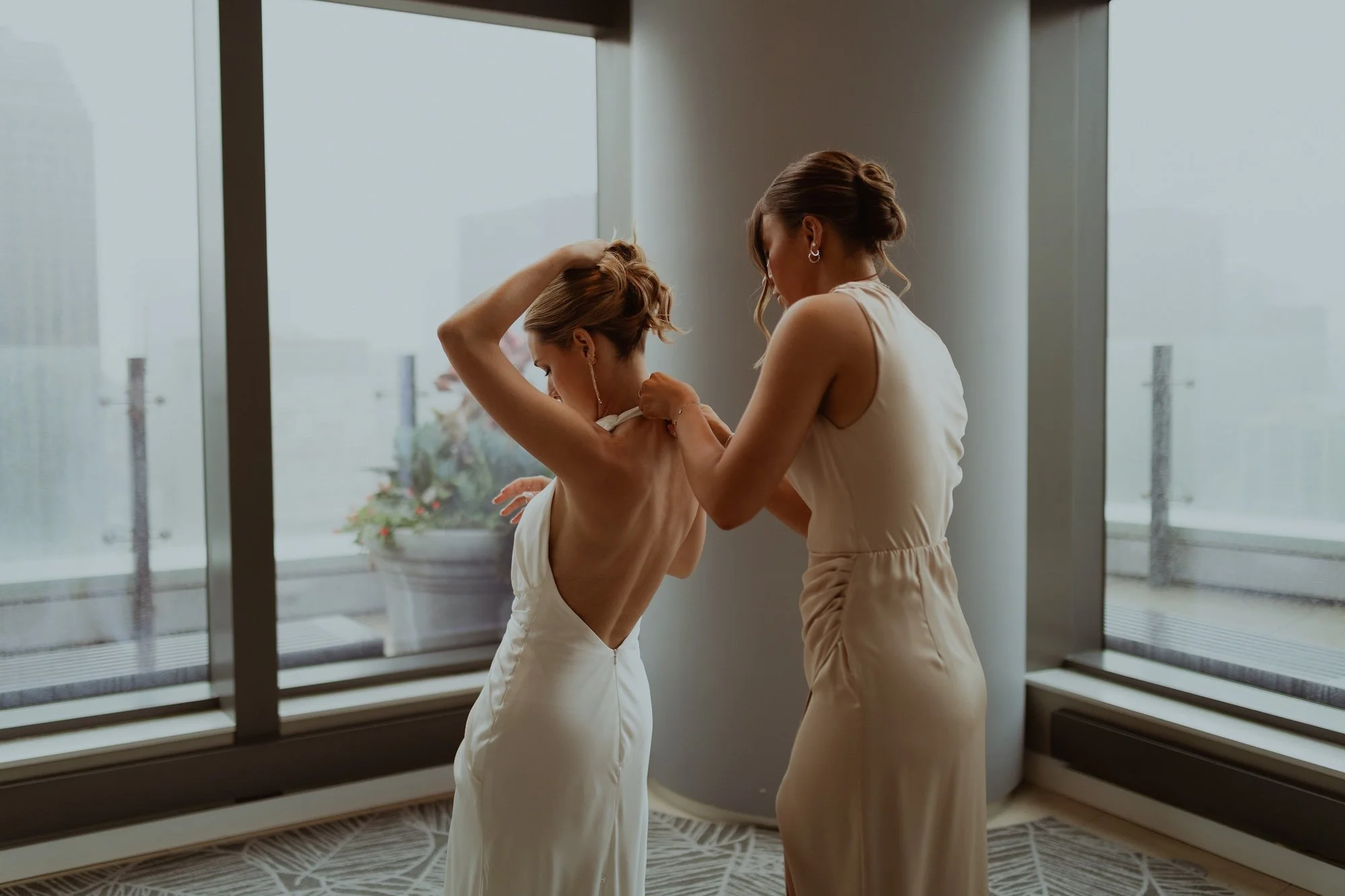 Two women in white dresses helping each other get ready, standing near large windows overlooking a city skyline, with a potted plant outside.