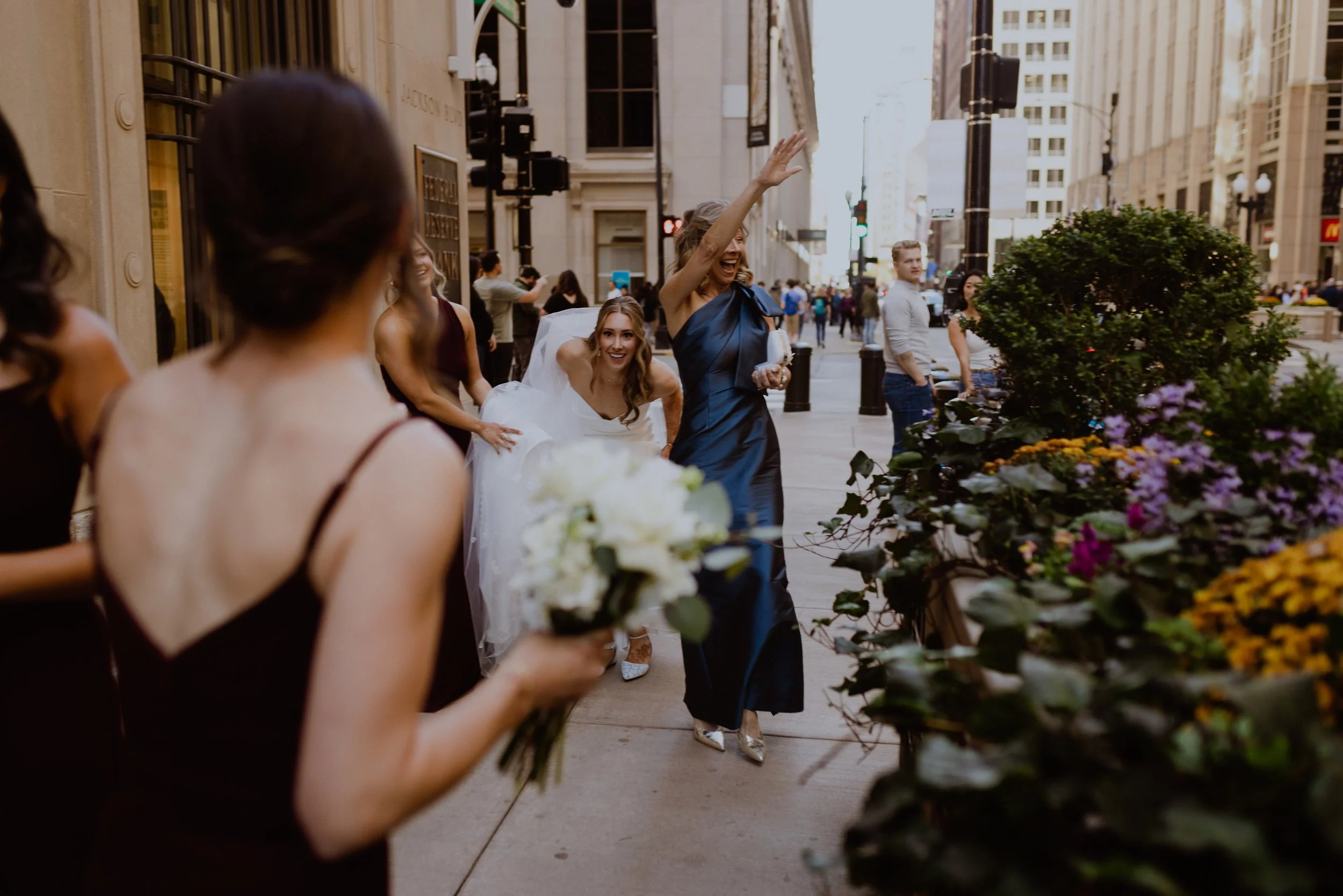 A woman in a dark blue dress(MOB) is waving on a city sidewalk during a wedding celebration, bride is crouching down to not be seen by the groom, surrounded by other women, some holding bouquets, with buildings and pedestrians in the background.