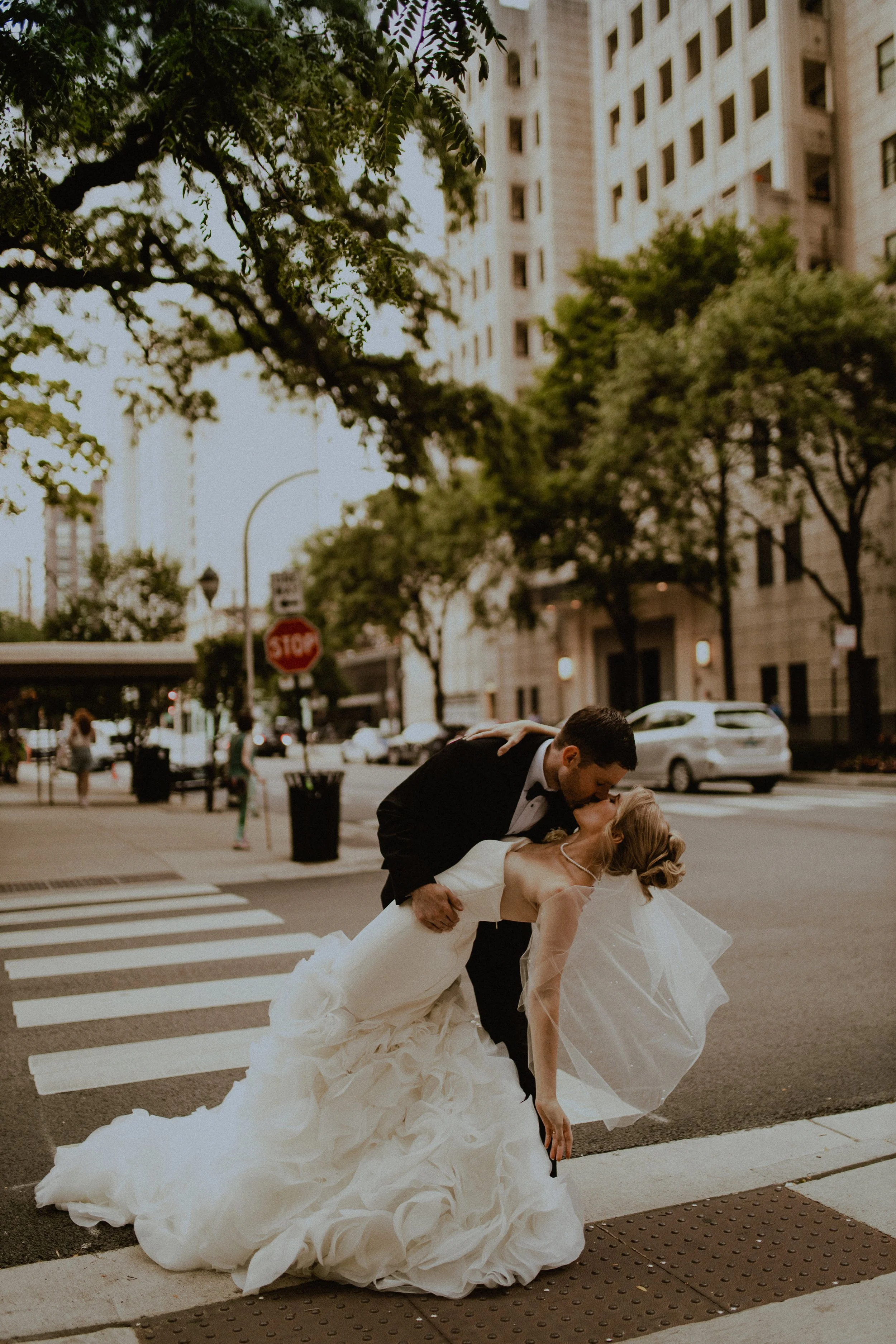 A bride and groom share a kiss on a city crosswalk, with the groom dipping the bride. She is wearing a white wedding gown and veil; he is in a black tuxedo. Tall buildings and trees are in the background.
