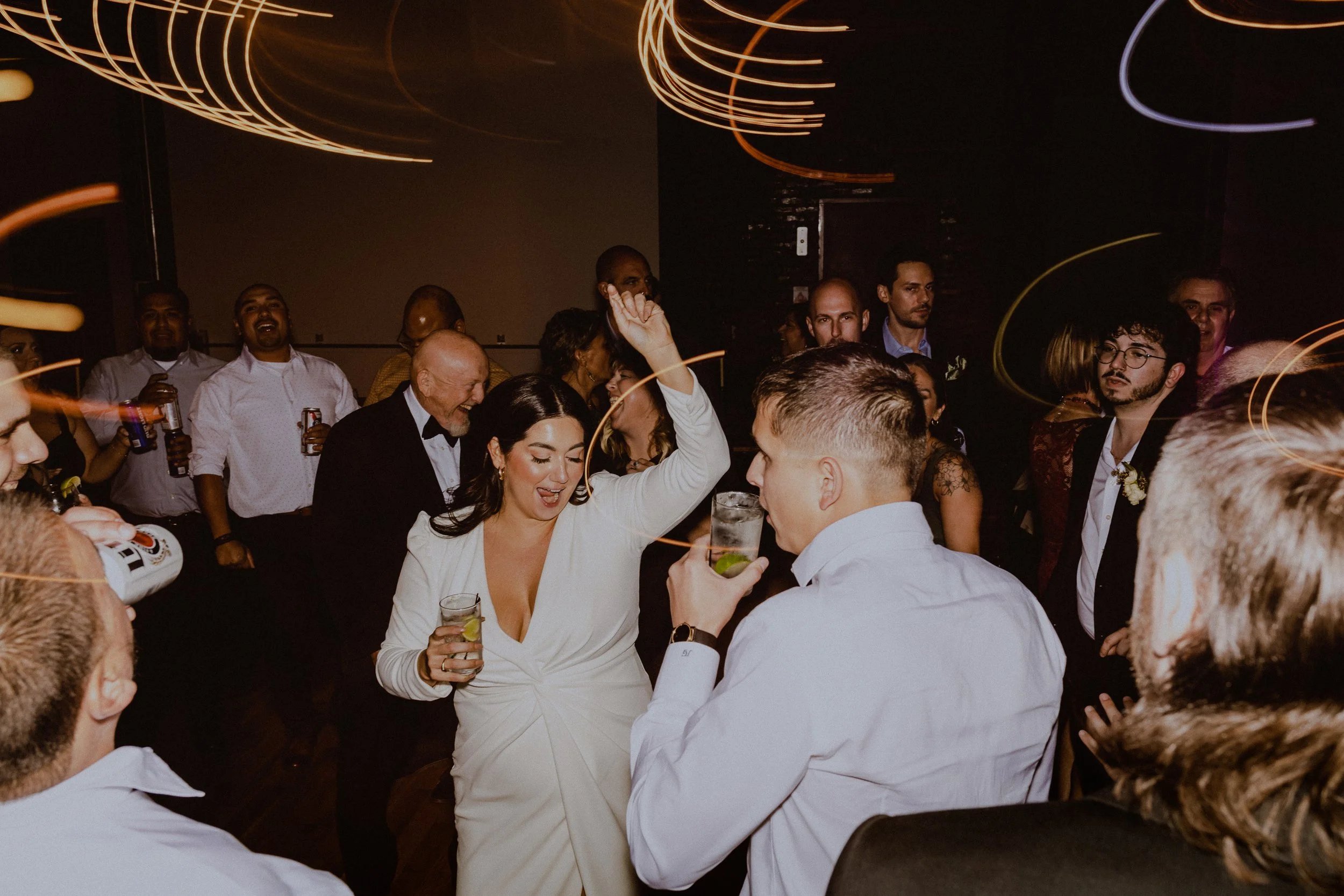 A woman in a white dress dancing and holding a drink at a lively party or celebration with many people surrounding her, some holding drinks and some dancing, in a dimly lit room with swirling light effects.