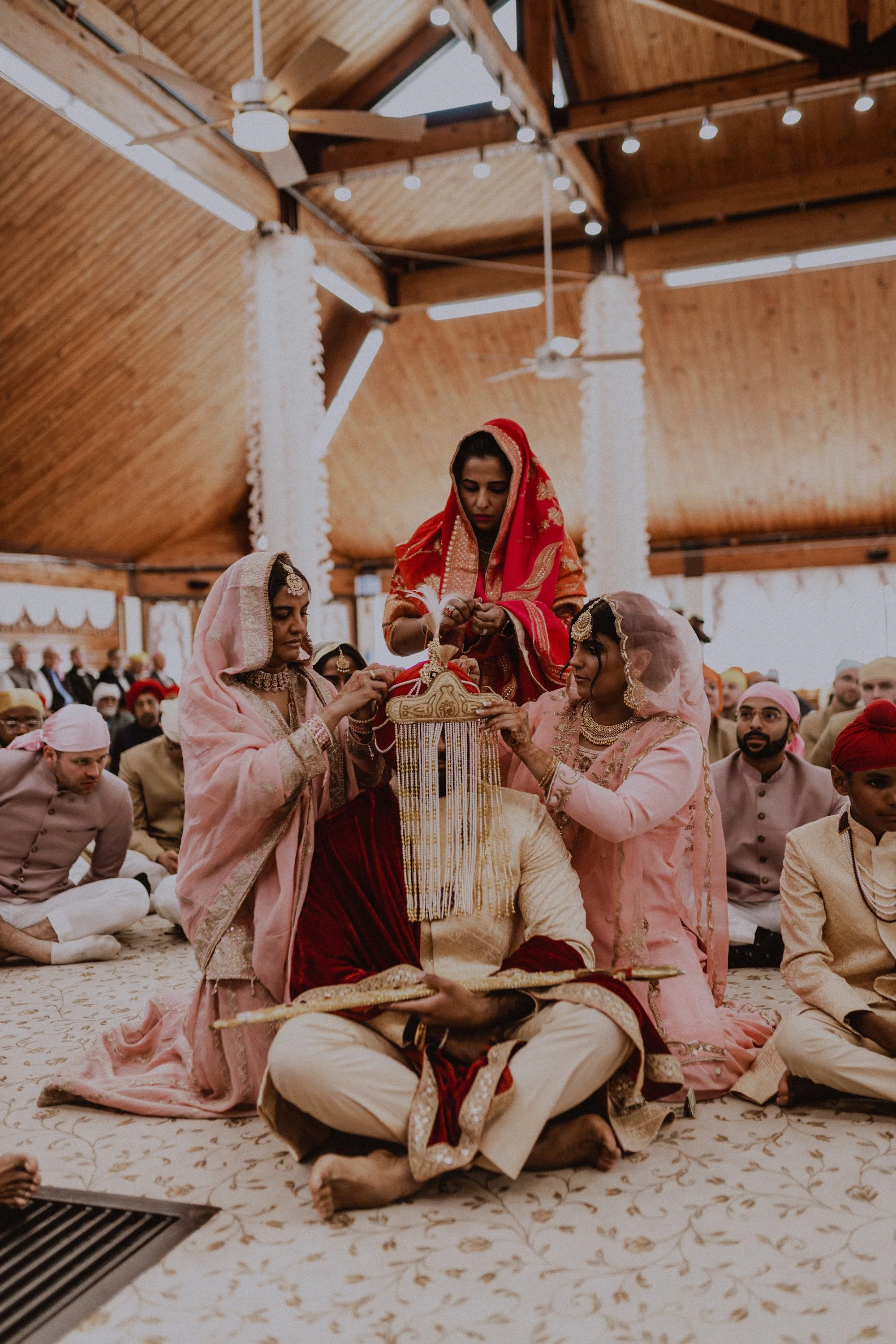 Indian wedding ceremony with women in traditional attire performing rituals around a seated groom holding a book, inside a decorated hall with wooden ceiling and string lights. Chicago documentary, colorful, candid wedding photos by ttolbphotog