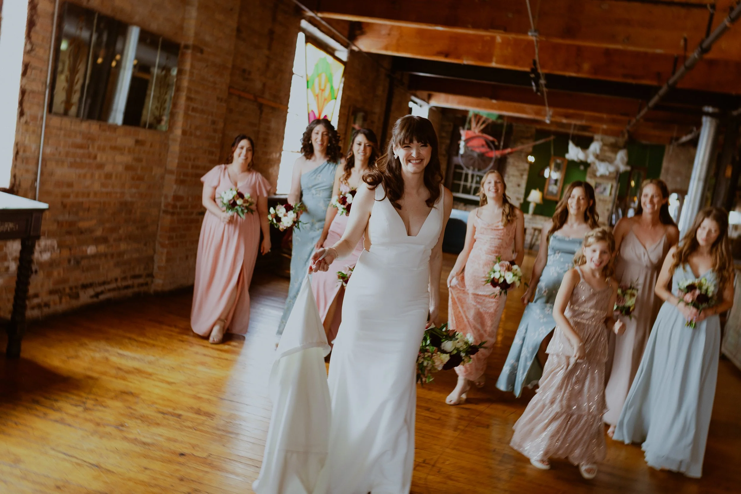 Bride in a white wedding dress walking into a room with bridesmaids and flower girls holding bouquets, in a rustic brick venue with wood floors and decorative elements at Salvage One Events