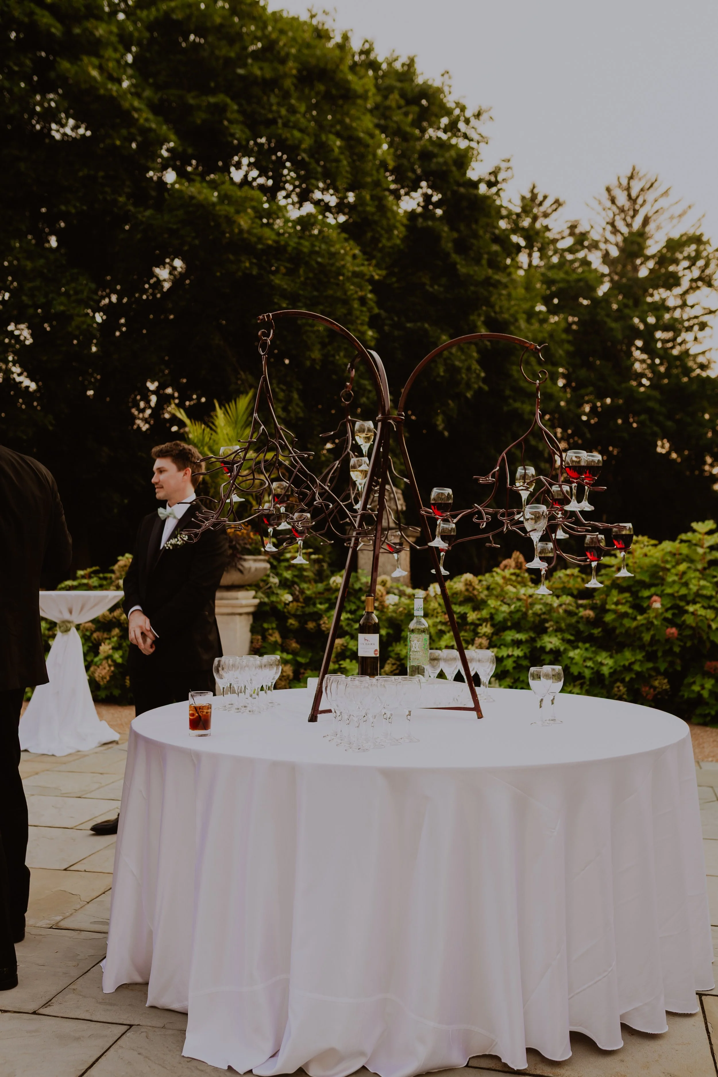 Outdoor event with a round table covered in a white tablecloth, decorated with glassware and bottles, and an ornate metal wine holder with glasses of red and white wine. Two men in tuxedos are visible in the background, in front of lush green trees a