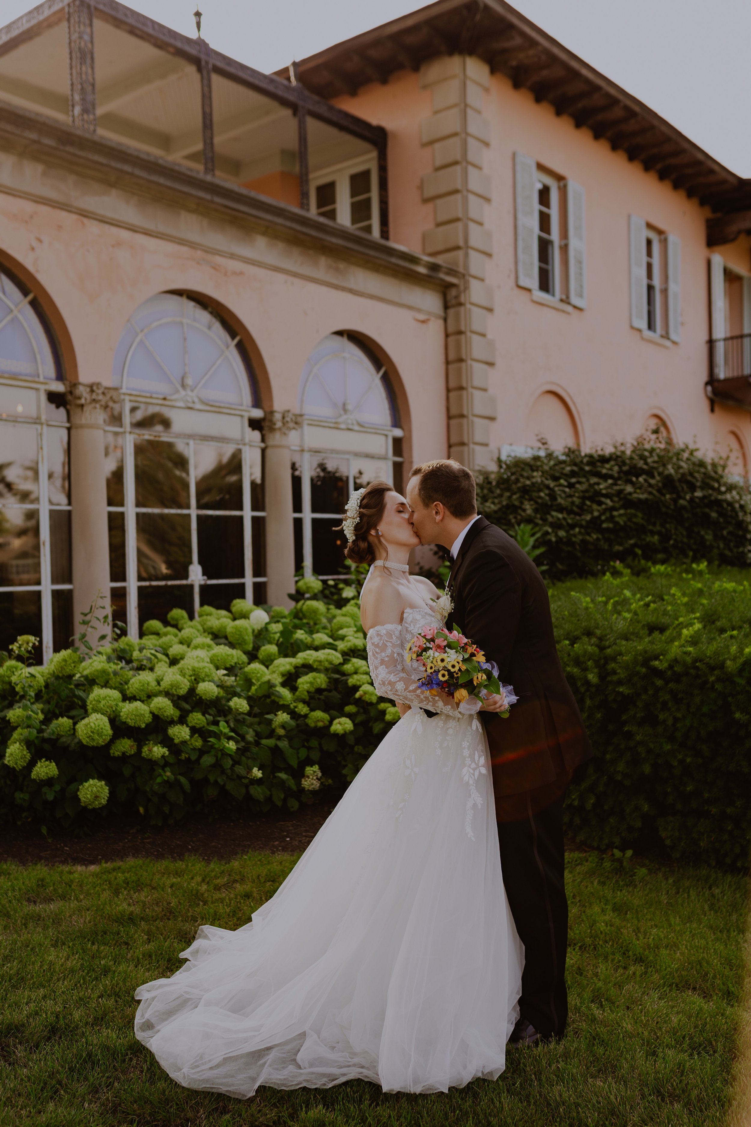 A bride and groom kissing outdoors in front of a building with pink walls and large arched windows, the bride holding a bouquet of flowers, surrounded by greenery and blooming bushes.
