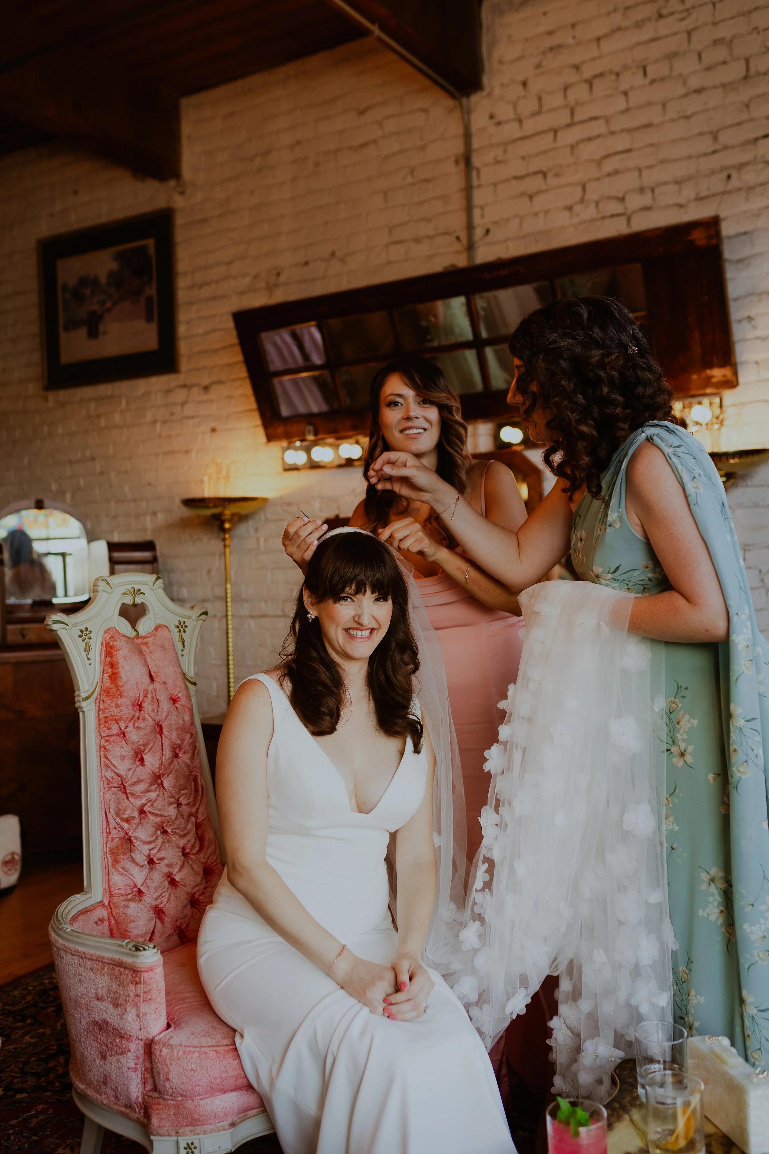 Salvage One Wedding Venue Chicago. Women sitting and standing at a social gathering, one in a white Weddinf dress sitting on a pink velvet chair, others in dresses, with a brick wall background, and drinks on a table nearby. Candid Wedding Photos