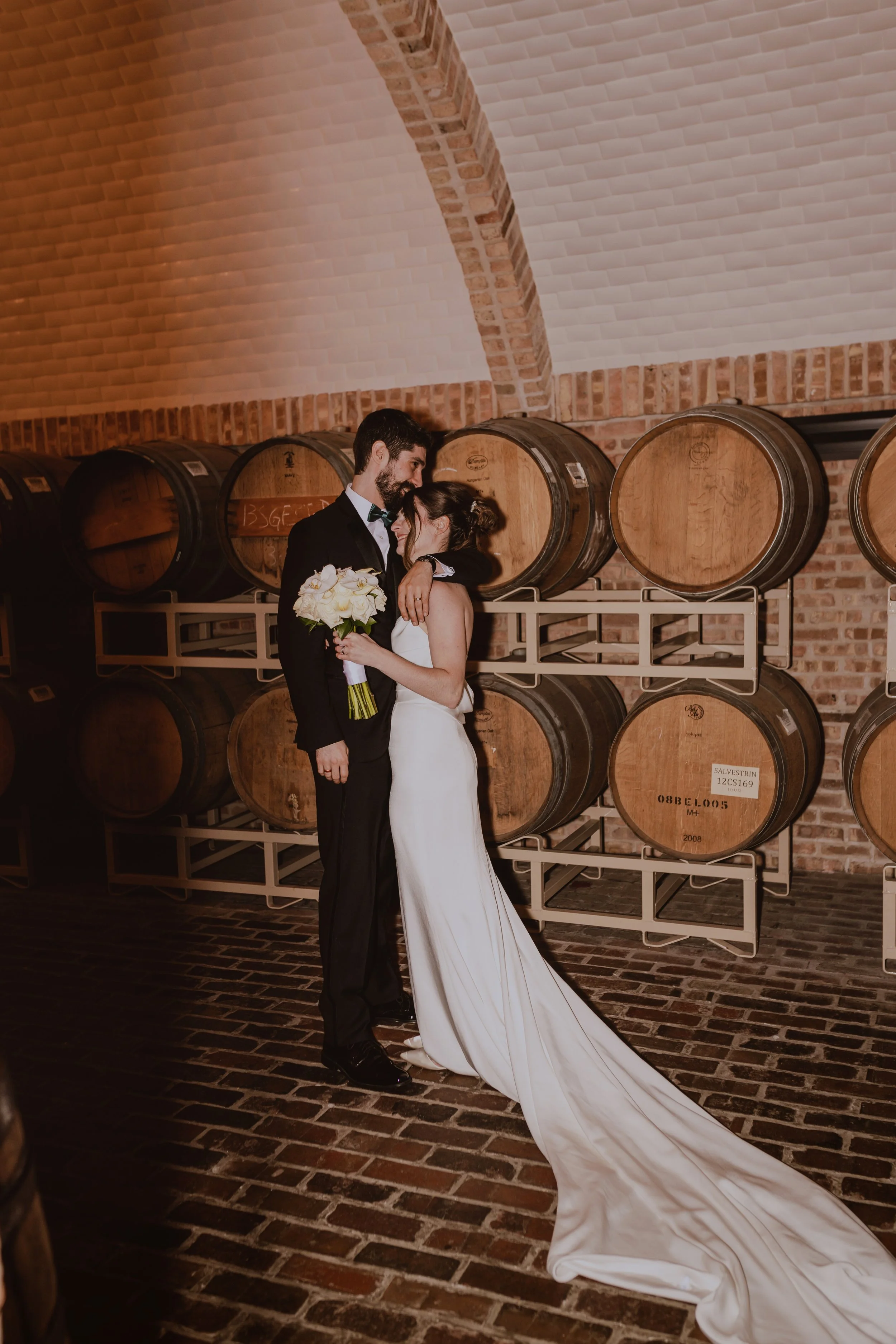 A newlywed couple sharing a romantic moment in a wine cellar, surrounded by wine barrels and brick walls.