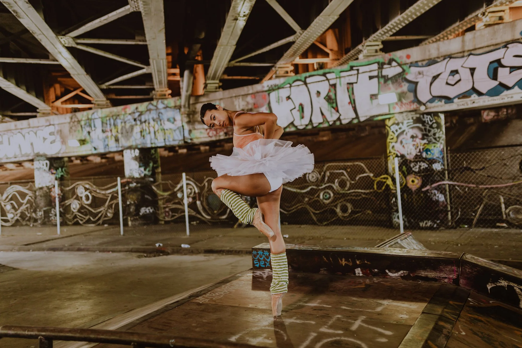 ballerina in white and green stripe leg warmers with white tutu and orange leotard on point in front of graffiti wall murals