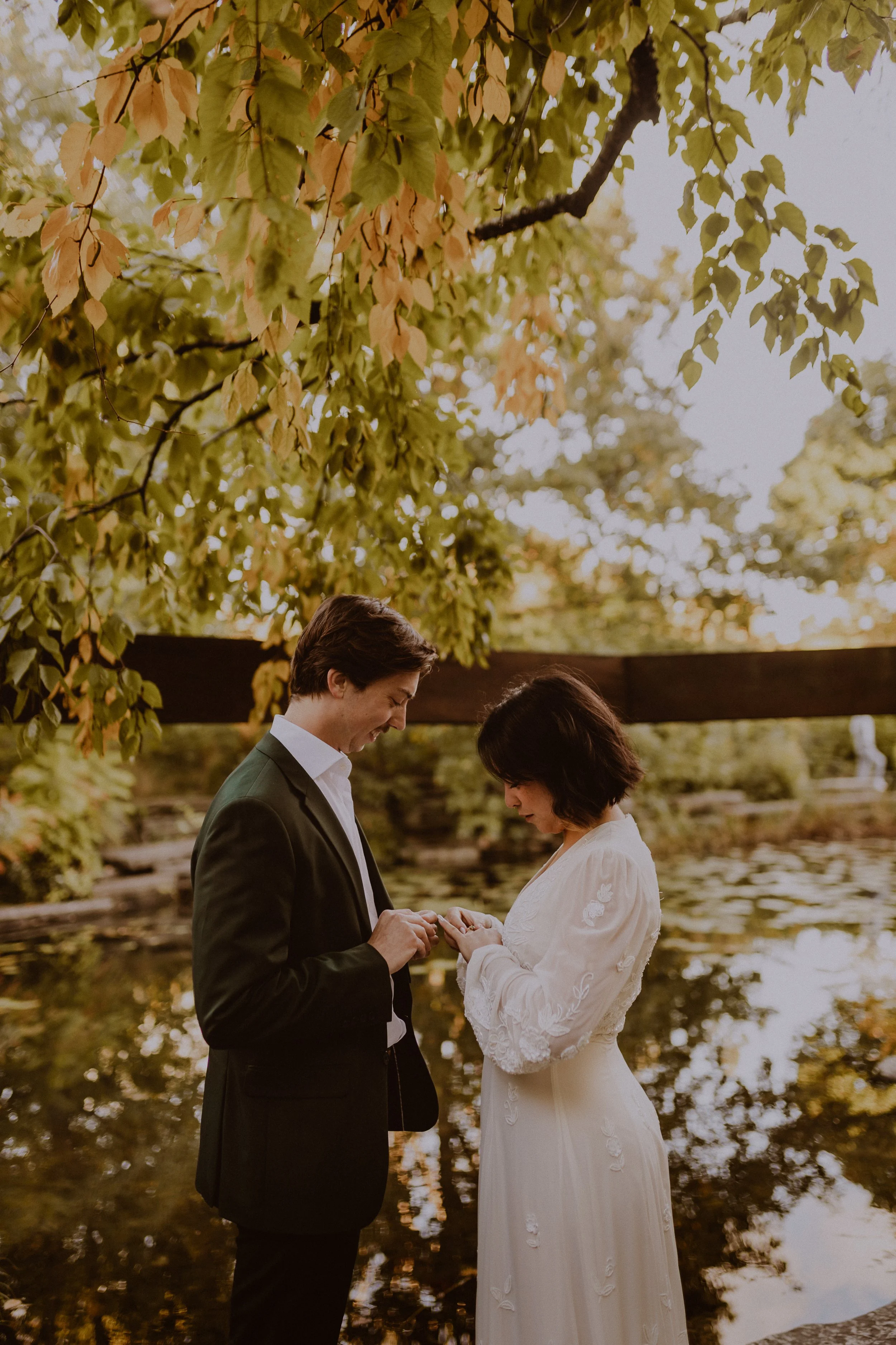 A couple dressed in wedding attire having a moment by a pond, with trees and a wooden bridge in the background.