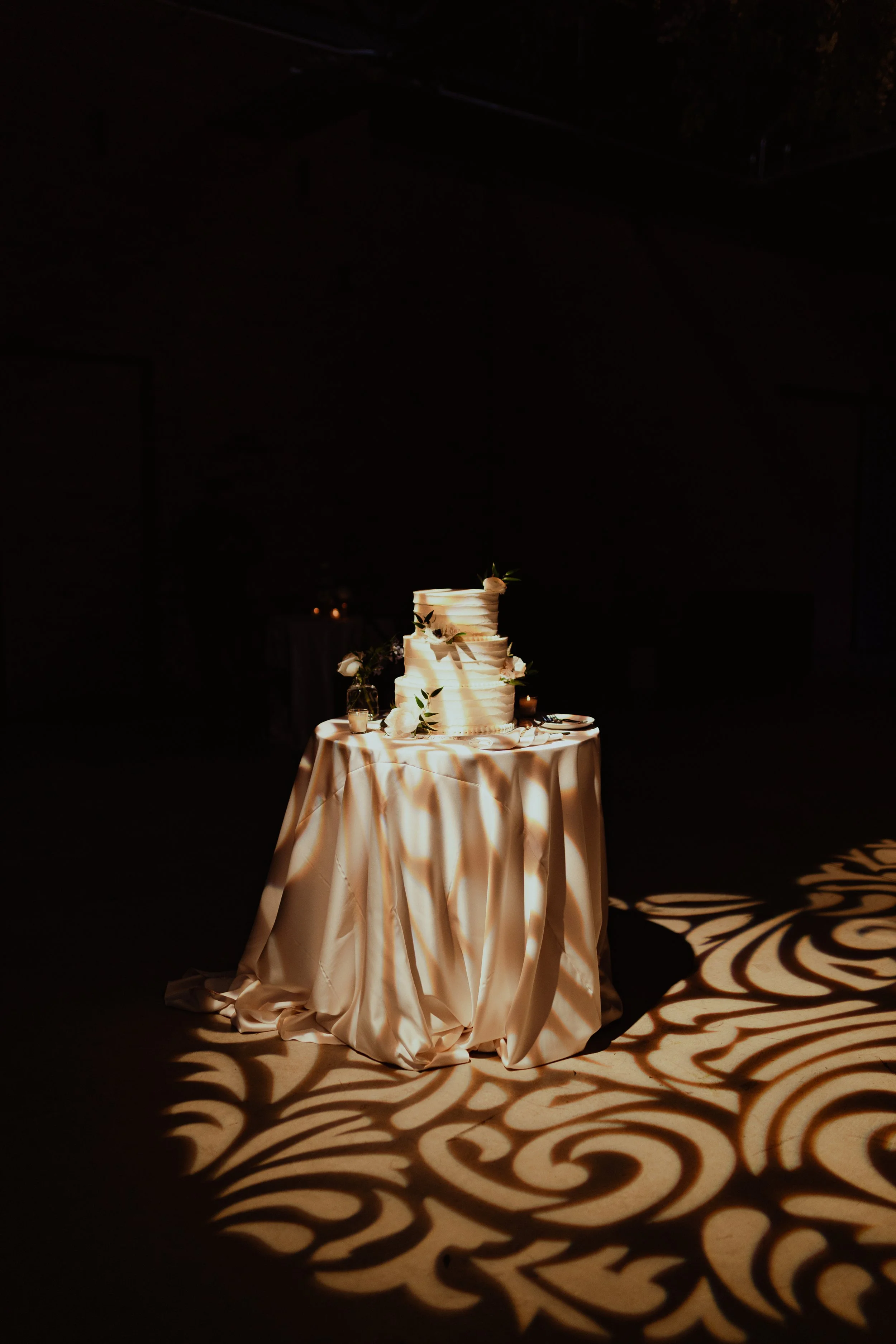 A four-tier wedding cake decorated with greenery and white flowers, sitting on a table with a white tablecloth. Dramatic lighting casts intricate shadows on the floor.