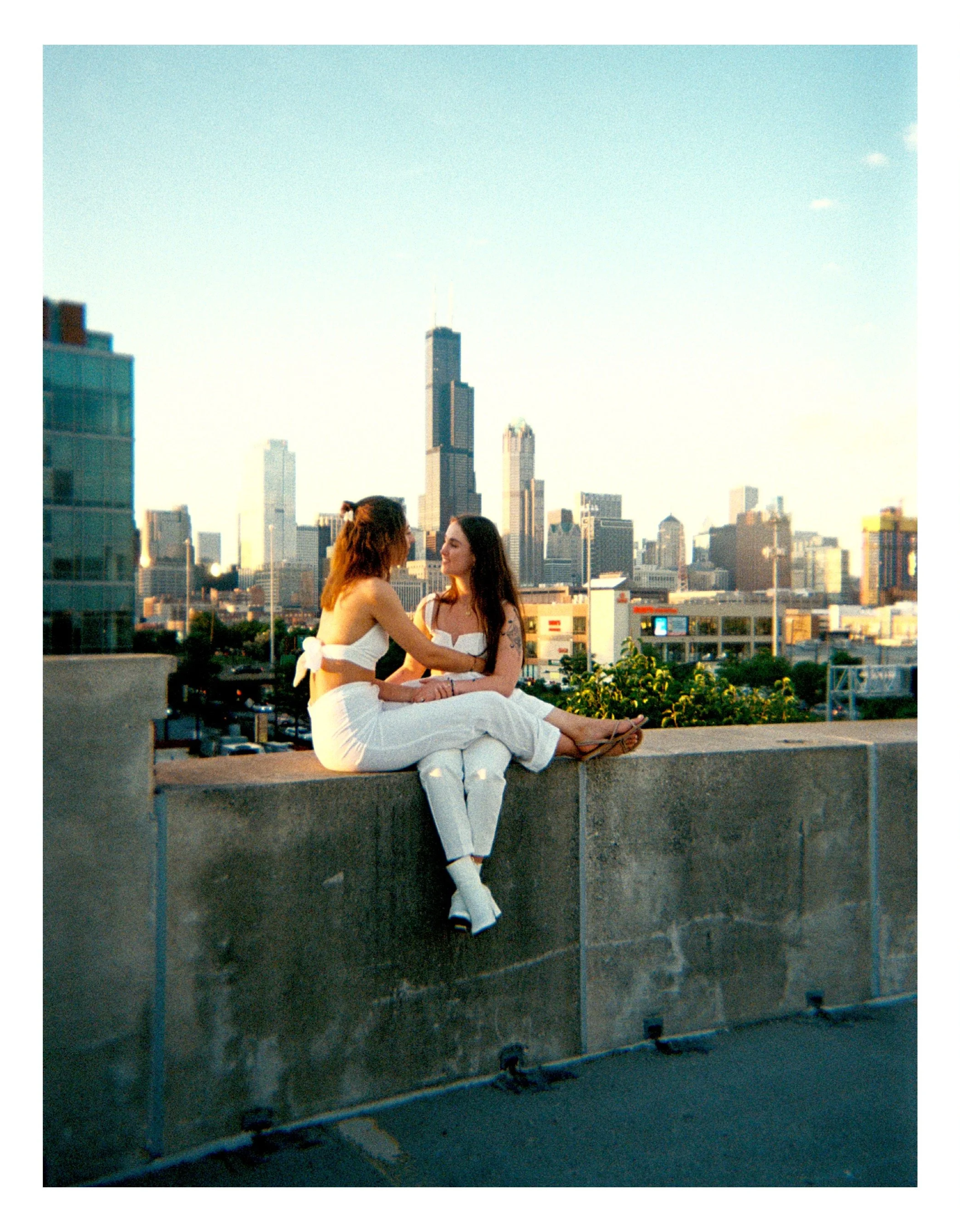 Double proposal for lesbian couple both in white suites, sitting on a concrete ledge on a rooftop, facing each other and holding hands, with a city skyline including the Willis Tower in the background during sunset.