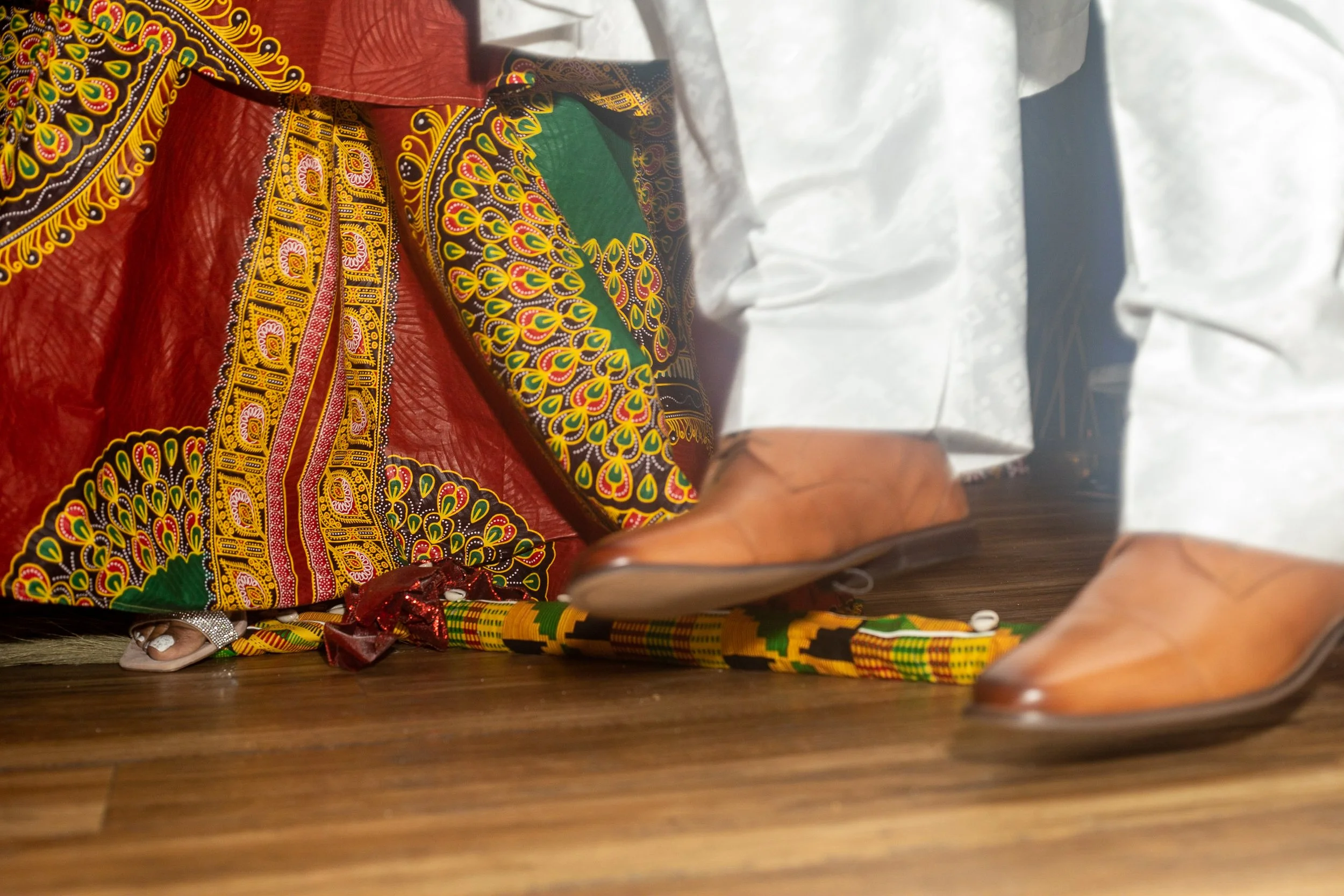 Feet of a person in traditional white attire with brown shoes walking over colorful beaded jewelry and vibrant patterned fabric on a wooden floor.