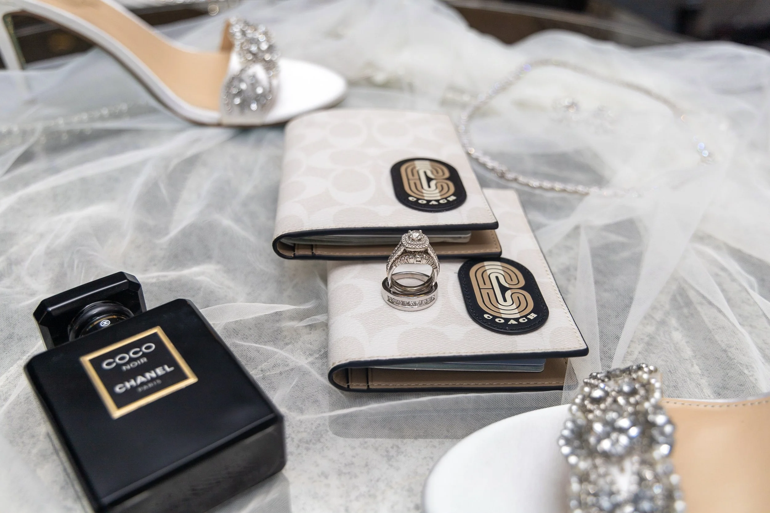 A collection of fashion items including two wallets, a diamond engagement ring, a Chanel COCO Noir perfume bottle, a pair of white high heels with rhinestones, a pearl and rhinestone jewelry piece, and a sheer white fabric background.
